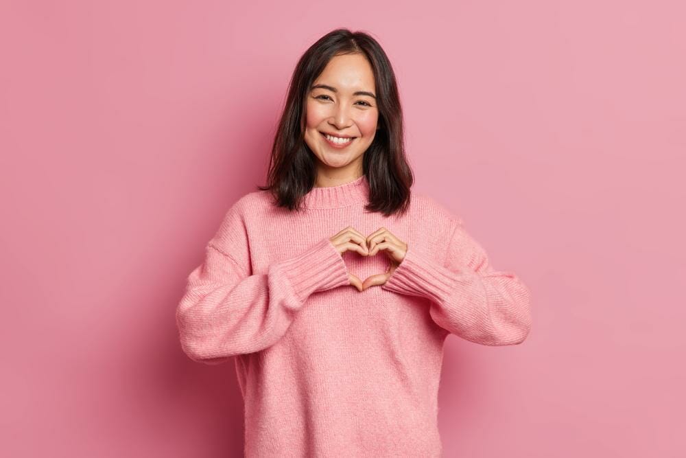 ピンクの背景に手でハートの形を作る韓国のアジア人女性。