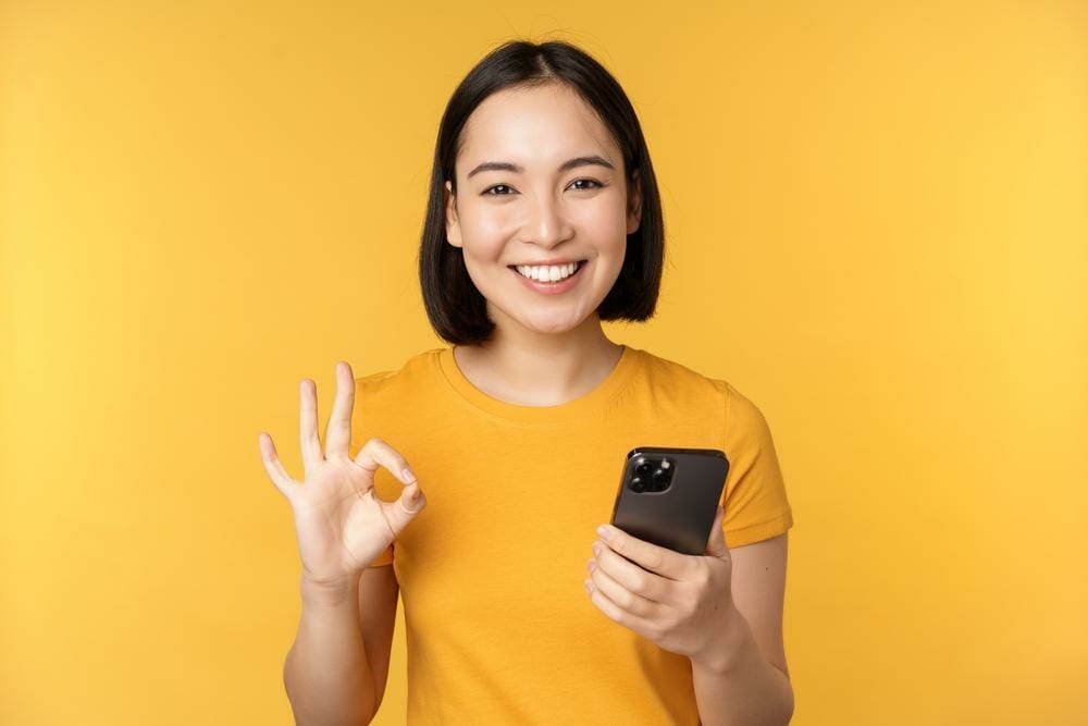黄色の背景にスマートフォンでOKのジェスチャーをする韓国の女性。