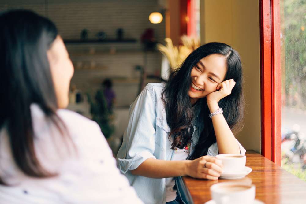 居心地の良いカフェの木のテーブルに2人の女性が座り、コーヒーを楽しんでいる。髪の長い女性の1人は微笑みながら、もう1人の女性を見つめながら手をかざしている。彼女たちは友好的な会話を交わしているようで、おそらく最近韓国を旅行した時の話をしているのだろう。