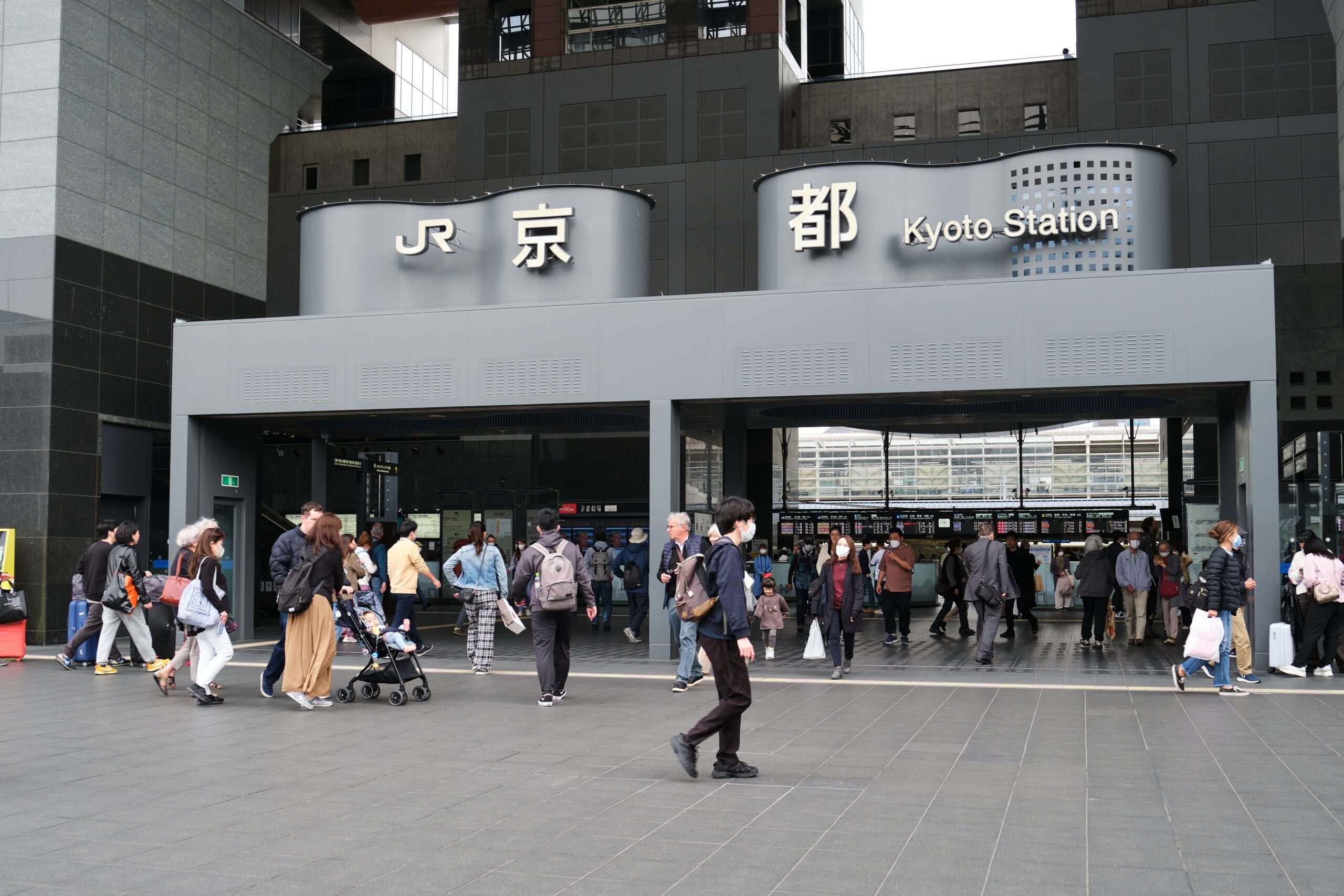 京都駅前を歩く人々。上には日本語と英語で「JR京都駅」と書かれた標識が見える。近くでは韓国語教室が韓国語学習に興味のある人を歓迎している。