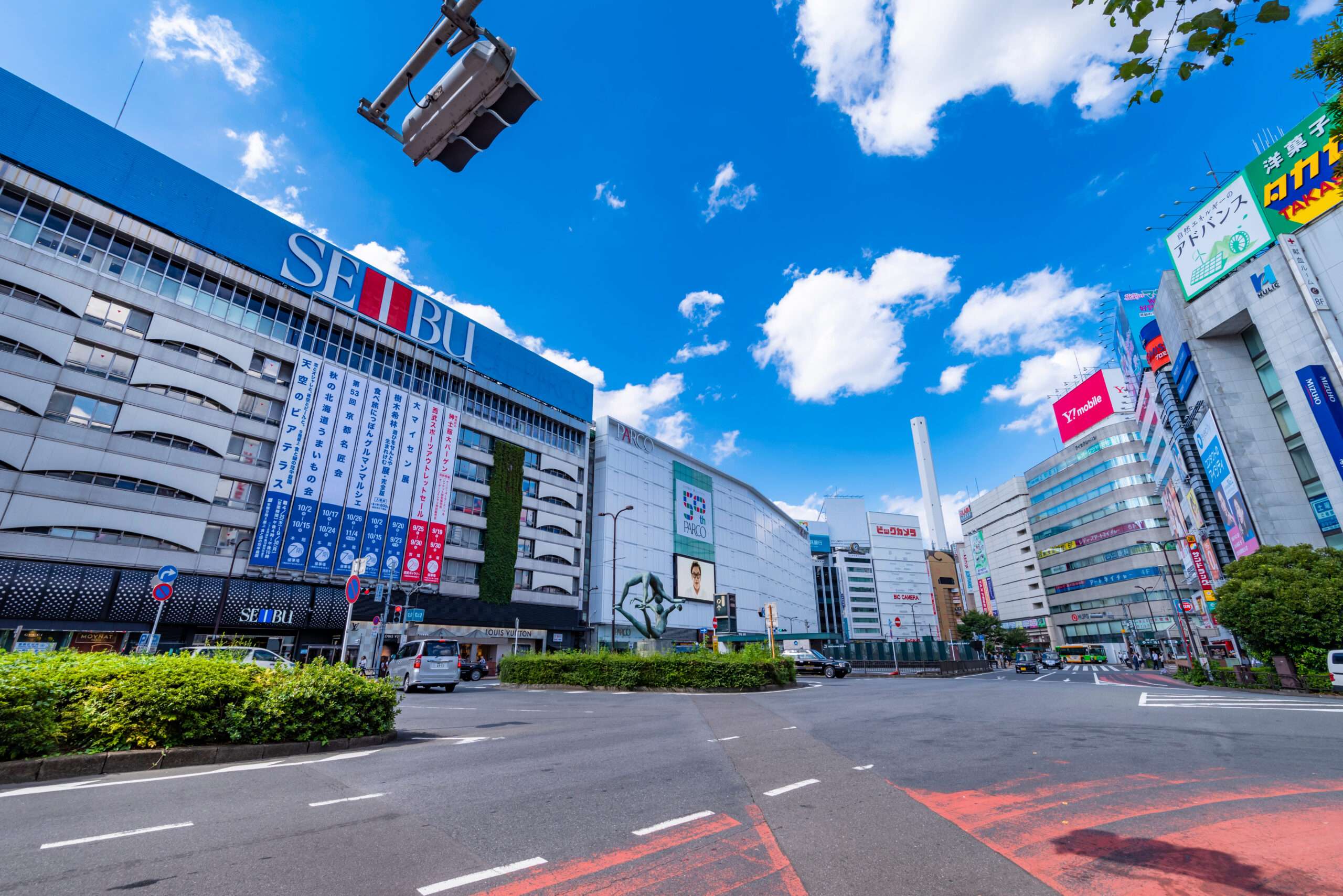 大型の西武百貨店を含む高層商業ビルが立ち並ぶ池袋の街並み。青空と散らばる雲が、活気に満ちた韓国の影響を思わせる活気あるエネルギーを響かせています。