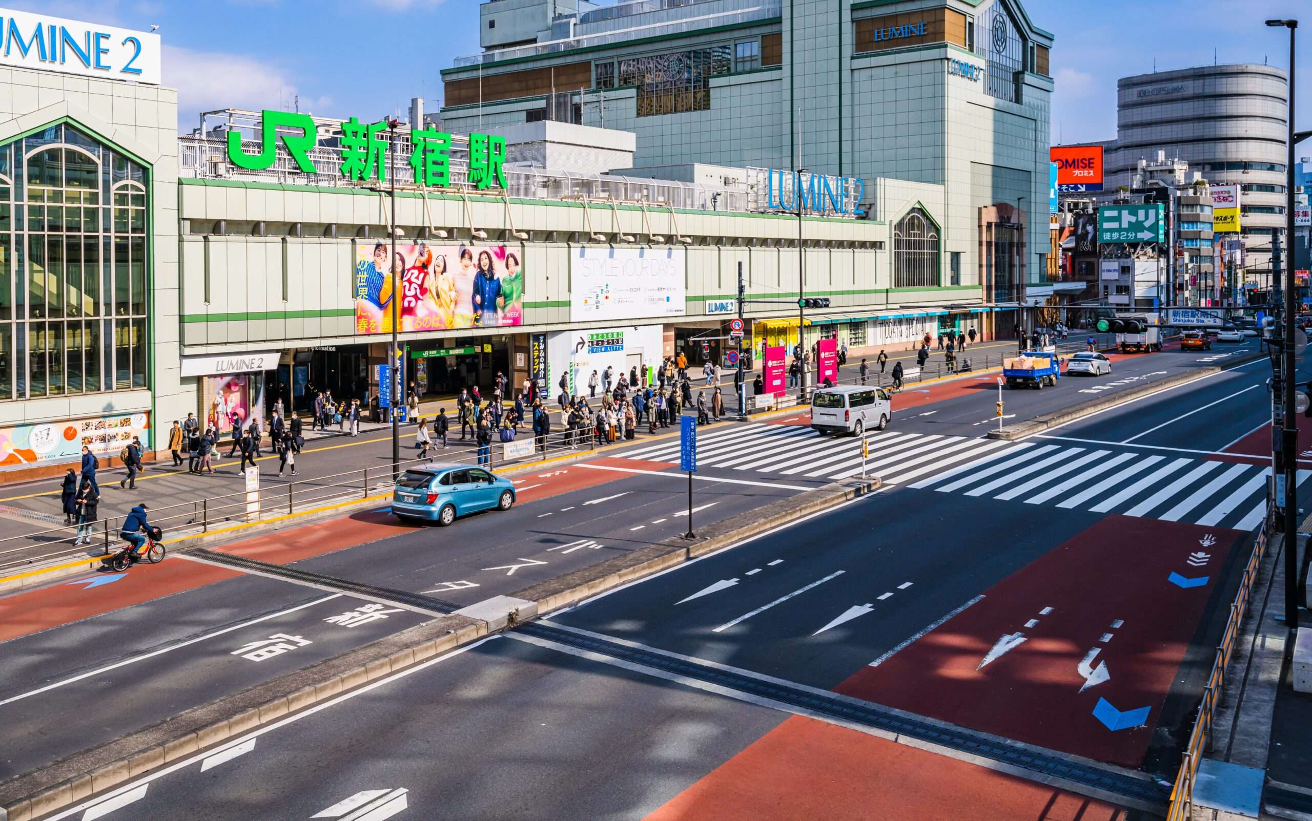 新宿の賑やかな市街地のストリートビュー。車、自転車、歩行者が「JR」と「ルミネ」と書かれた大きな建物の近くを行き交い、韓国語を話す人々の活発な会話が都市生活の活気あるタペストリーに彩りを添えています。