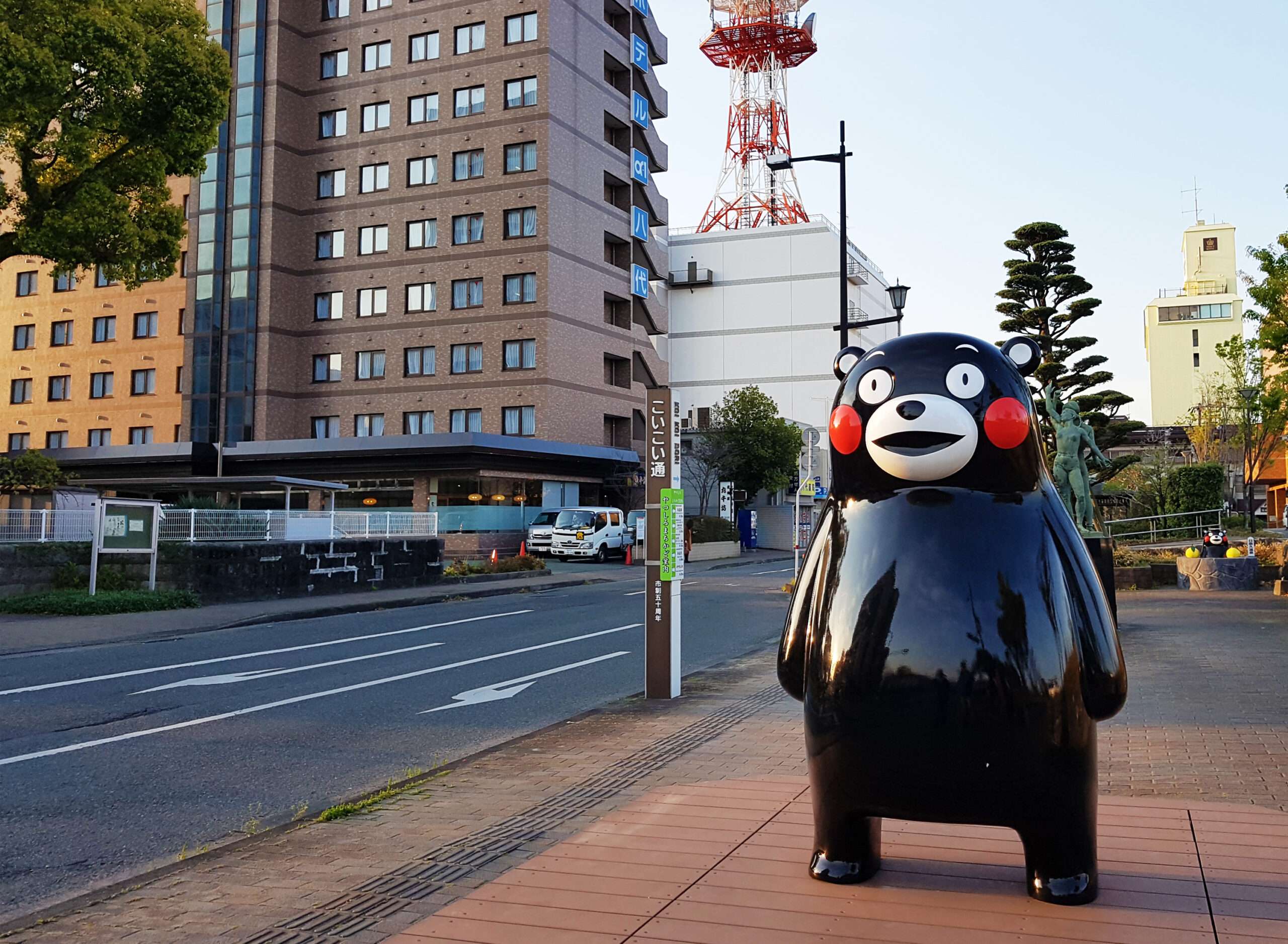 熊本の歩道に頬が赤い大きなツキノワグマの像が立っている。背景には高層ビル、道路、木々があります。近くでは韓国語教室に人々が集まり、シーンに文化的な活気を与えています。