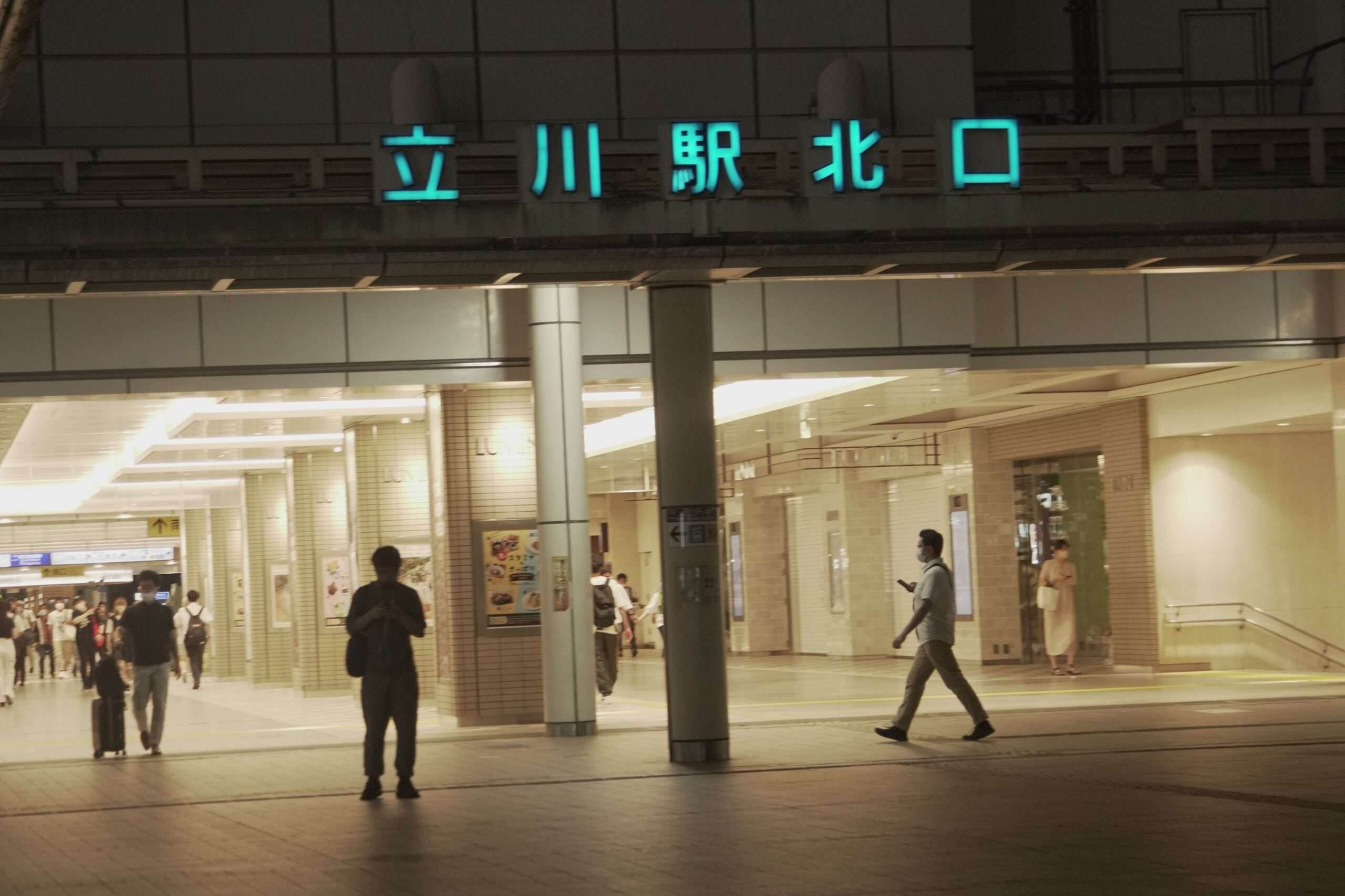 夜、立川駅の入り口付近を歩く人々。中には携帯電話に夢中になっている人もいる。駅のネオンサインが上に輝き、近くにある韓国語教室の広告が目を引く。