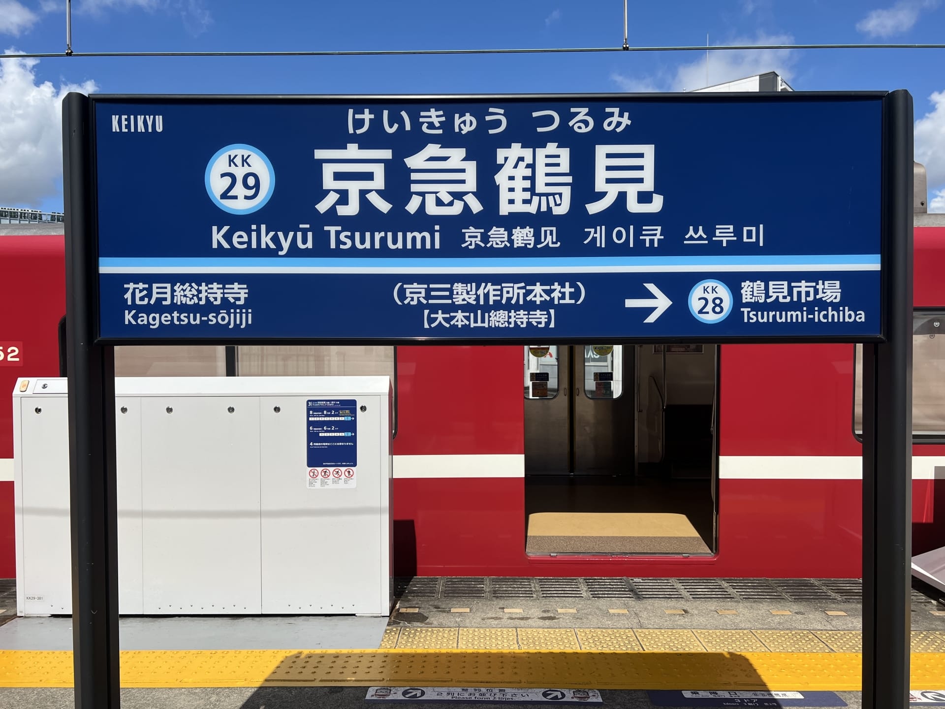 京急鶴見駅の看板。青い空の下、背景にホームと赤い電車が見えます。韓国語学習におすすめの教室からほど近い場所にあります。.