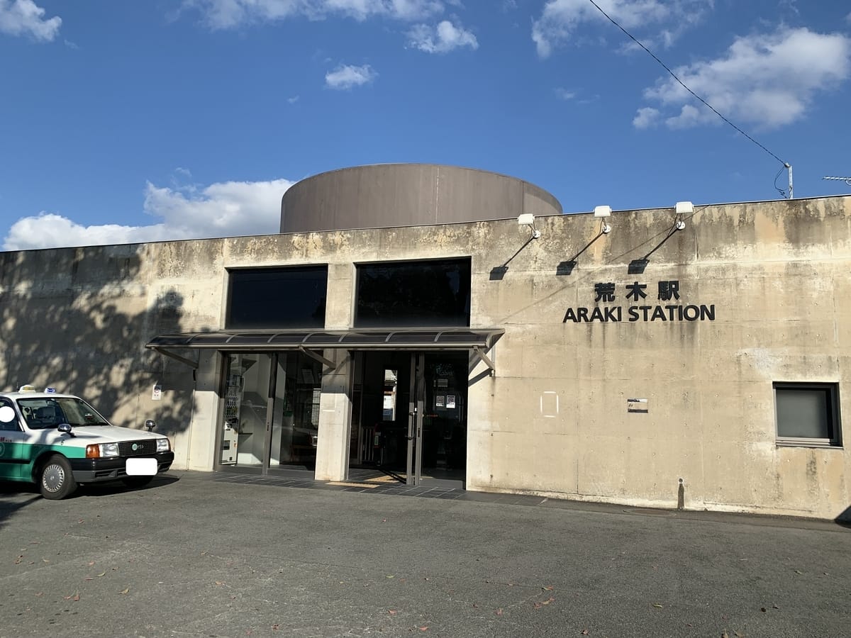 小さな近代的なコンクリートの建物に、英語と日本語で「ARAKI STATION」と書かれた看板。左側に緑のタクシーが停まっている。.
