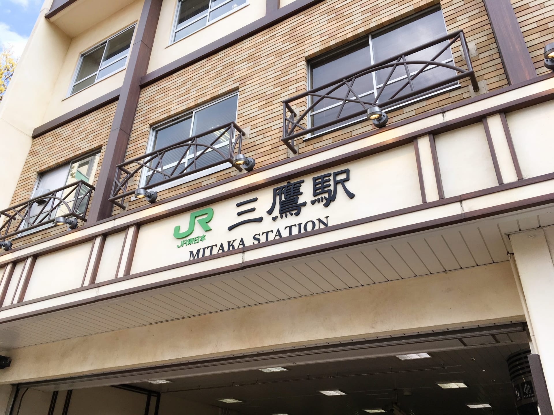 三鷹駅の入り口には日本語と英語の標識があり、木造の建物の外観にある象徴的な JR のロゴの下に、近くにある韓国語教室マンツーマンの詳細も表示されています。.