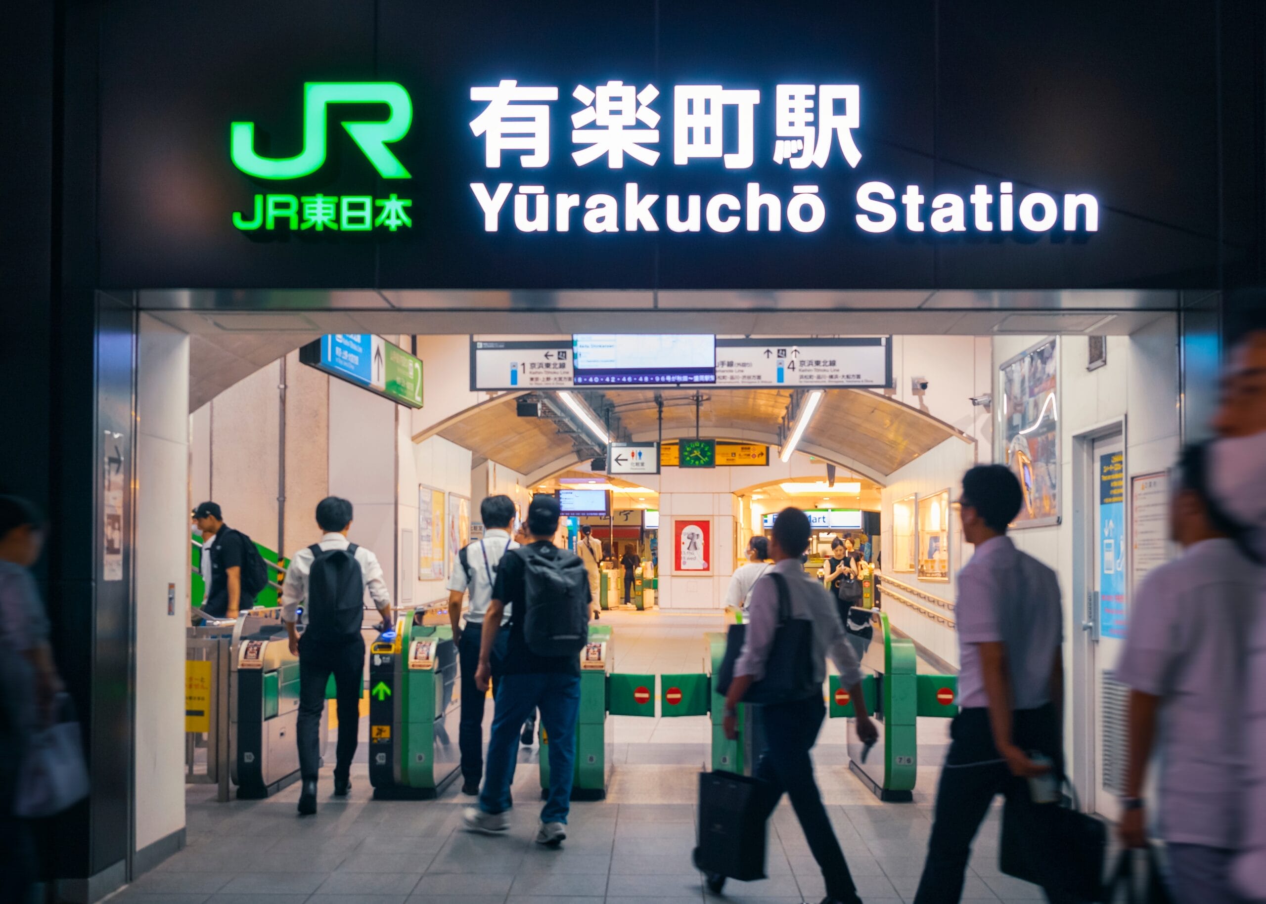 有楽町駅の改札を通る人々。上の駅名標は日本語とローマ字が併記されて点灯しています。近くの韓国語教室マンツーマンでは、マンツーマンで韓国語を学びたい人向けに個別の言語レッスンを提供しています。.