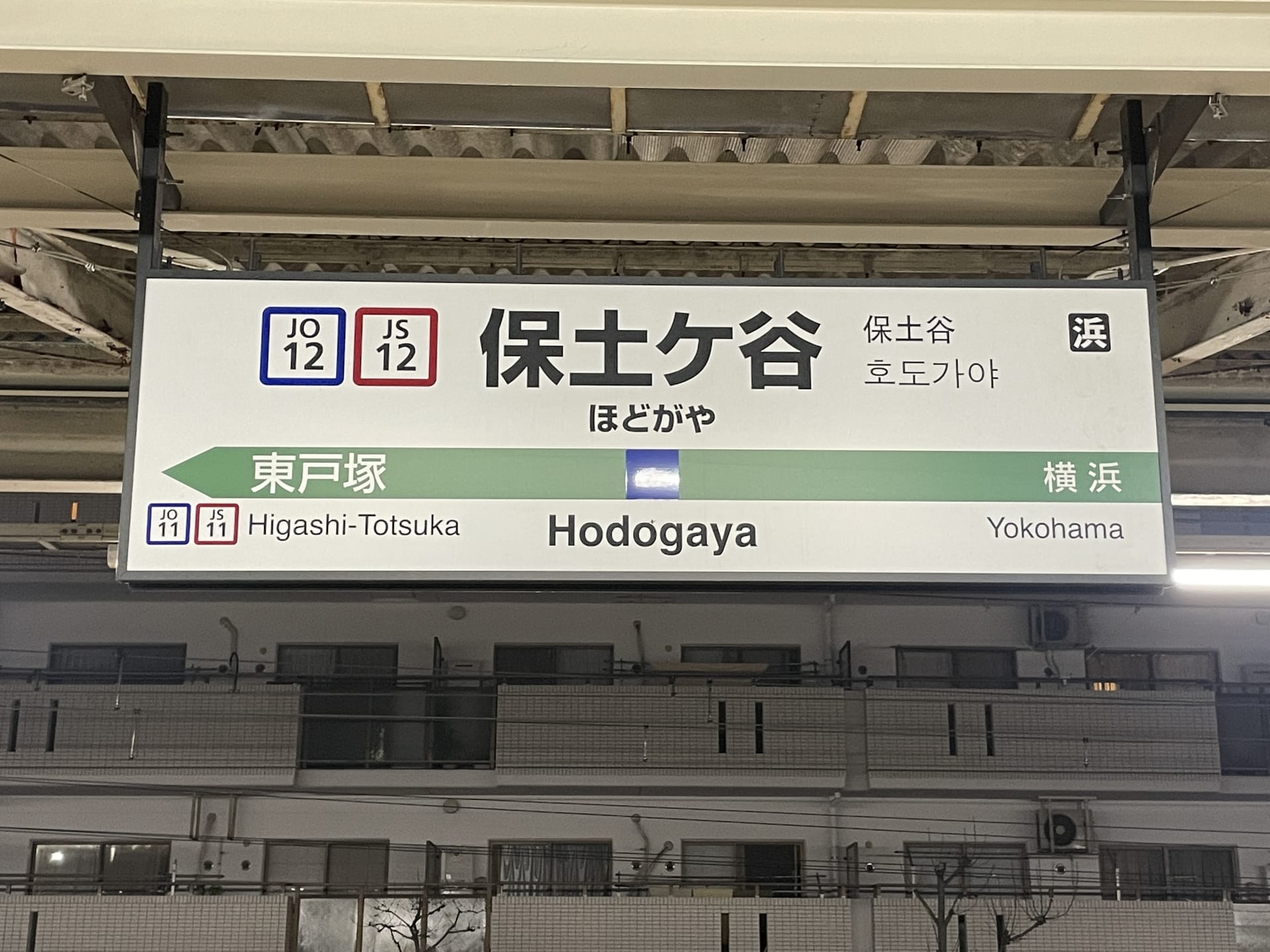 保土ヶ谷の駅名標は日本語と英語で表示され、東戸塚と横浜への方向が記載されており、駅コードは JO12 と JS12 です。韓国語教室マンツーマンやその他の目的地へ向かう通勤者に最適です。.
