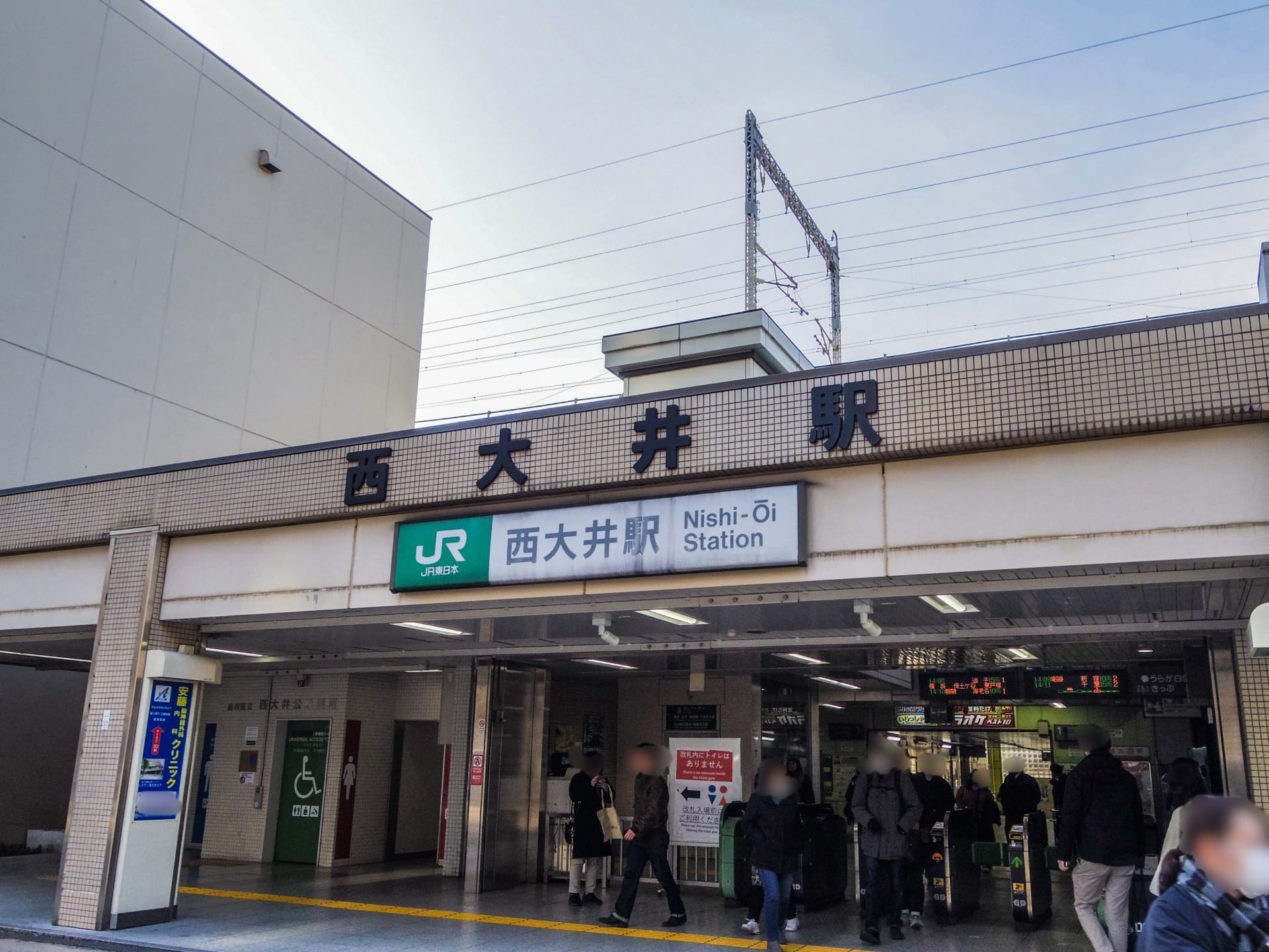 西大井駅の入口には日本語と英語の標識が設置されています。人々が行き交い、近くの韓国語教室マンツーマンへ向かう人もおり、都市部の活動と教育活動が融合しています。.
