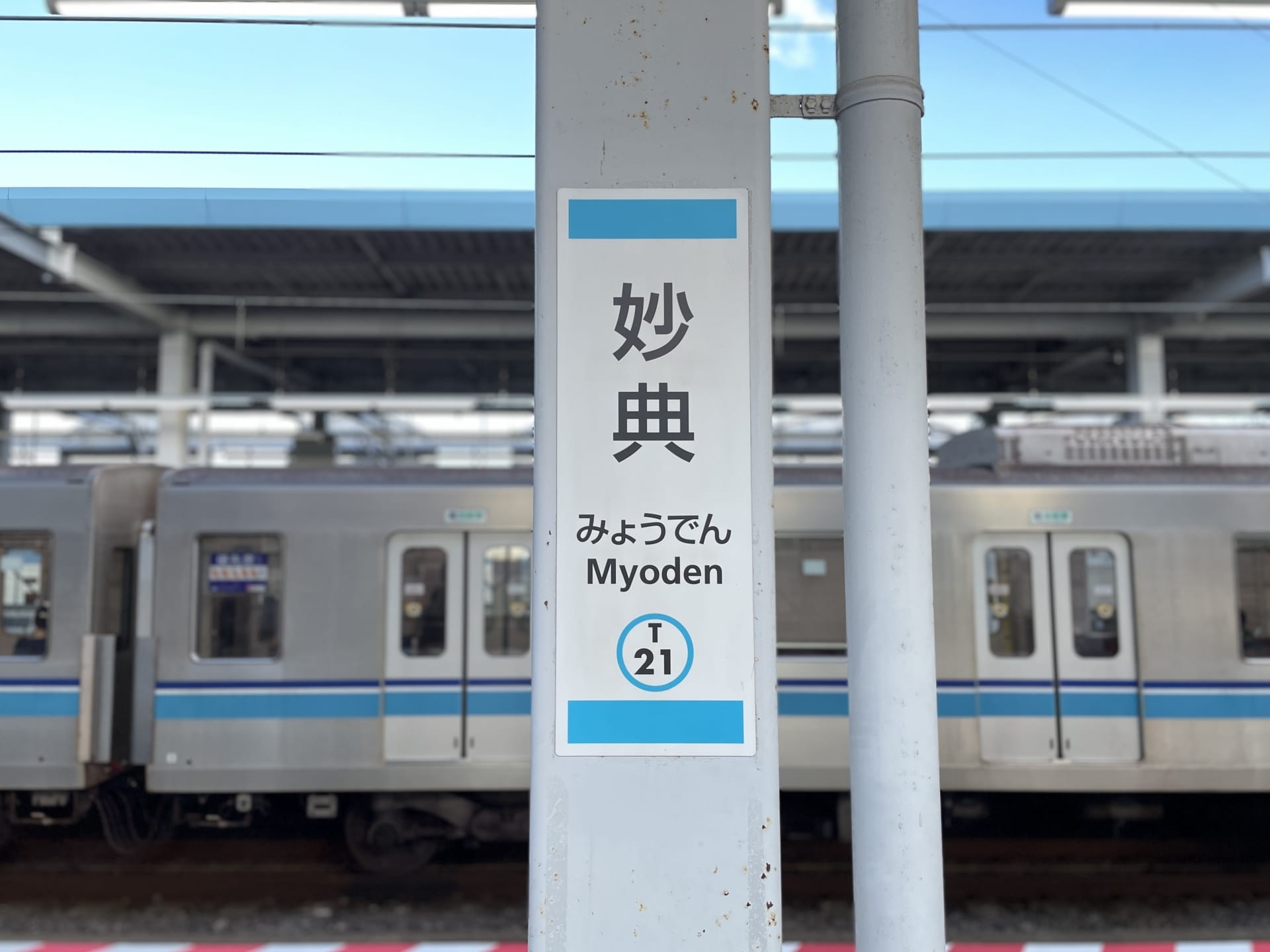 妙典駅の標識。背景には電車が写っており、駅名「妙典」が日本語、韓国語、英語で表示され、駅番号はT21となっている。.