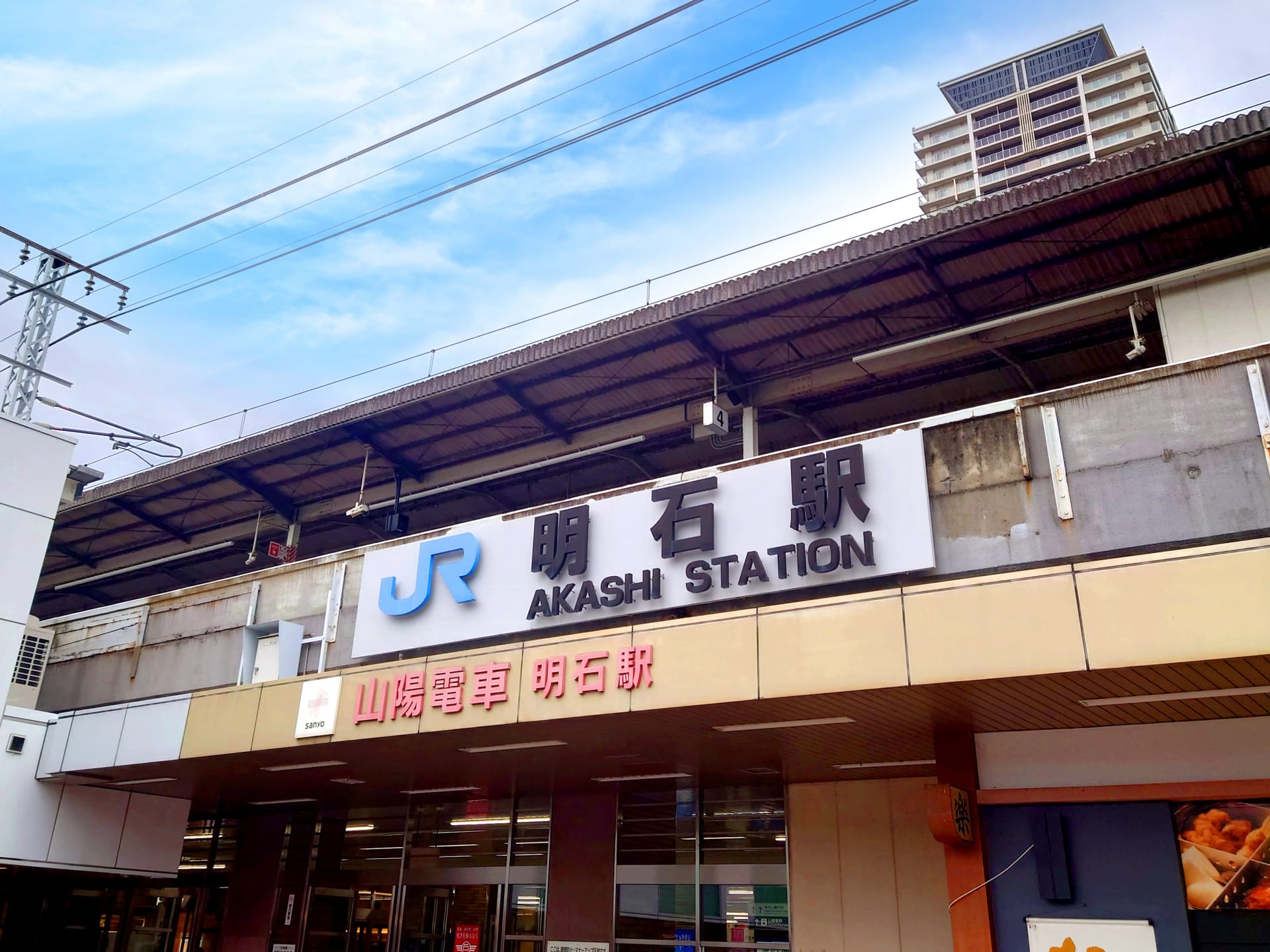 明石駅の正面図。JRのロゴと日本語、英語、韓国語の駅名標が掲げられています。建物は曇り空の下、明石の文化の融合を体現しています。.