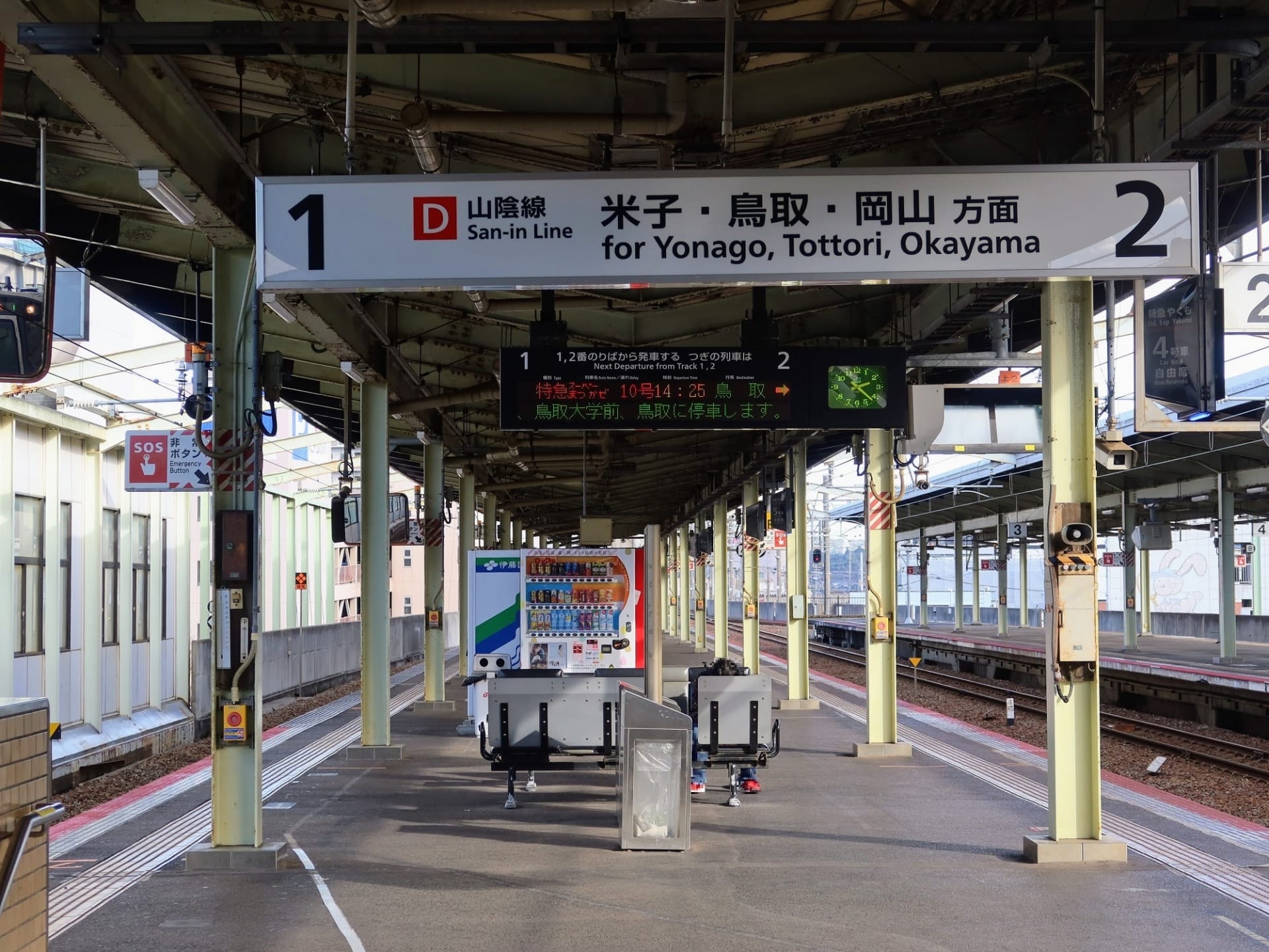 2 本の線路がある空のホームには、米子、鳥取、岡山、松江・島根を表示する頭上標識が設置されています。構造梁と照明で保護されており、便利な自動販売機も設置されています。.