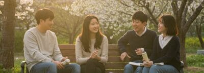 桜が咲いている公園のベンチに4人が座り、日差しの中で話したり笑ったりしています。.