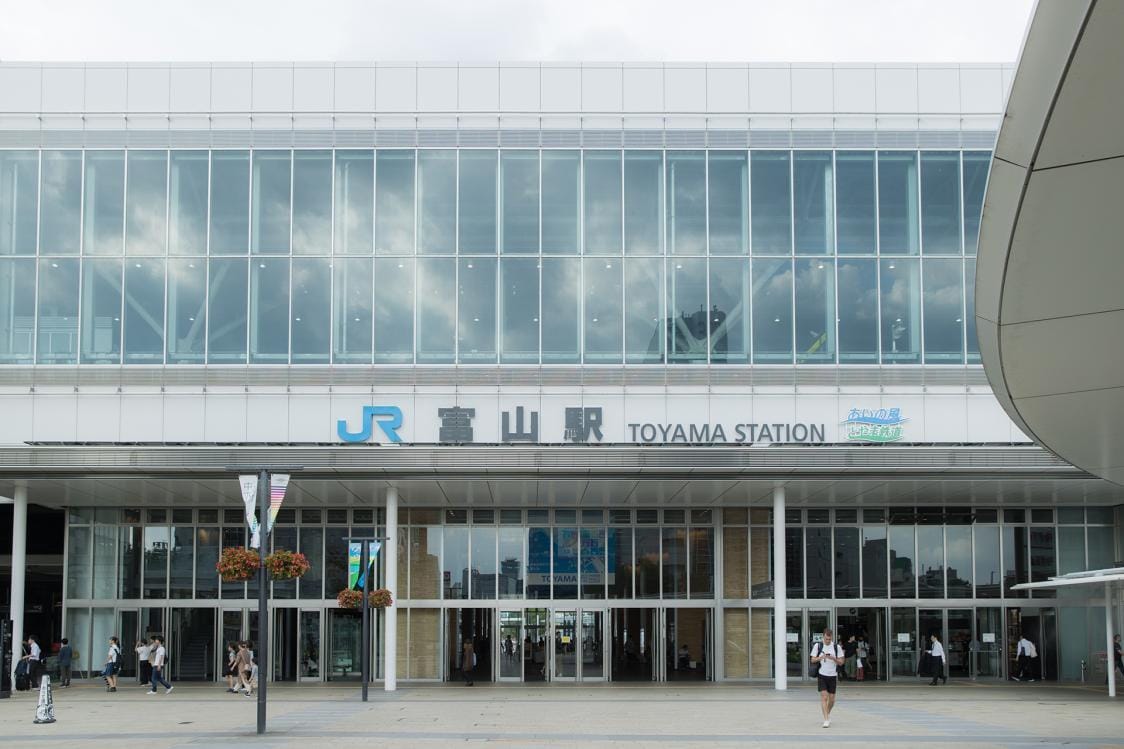 日本の富山駅の正面図。大きなガラス窓、見える看板、入り口の外を歩く数人の人々。.