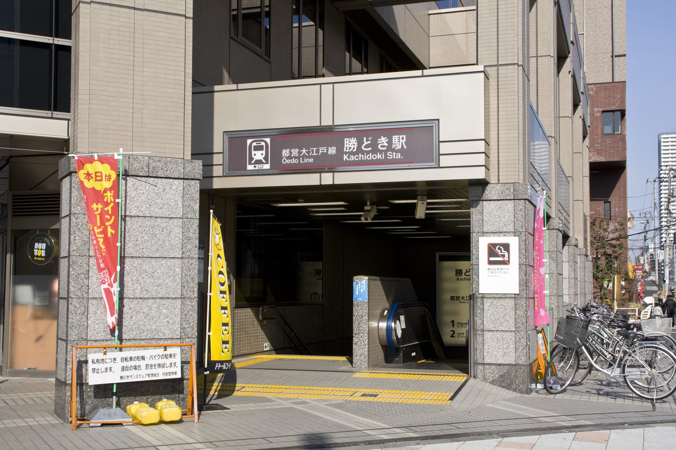 都営大江戸線勝どき駅の入り口。日本語の看板、エスカレーター、自転車、そして建物の外にはカラフルな横断幕。.