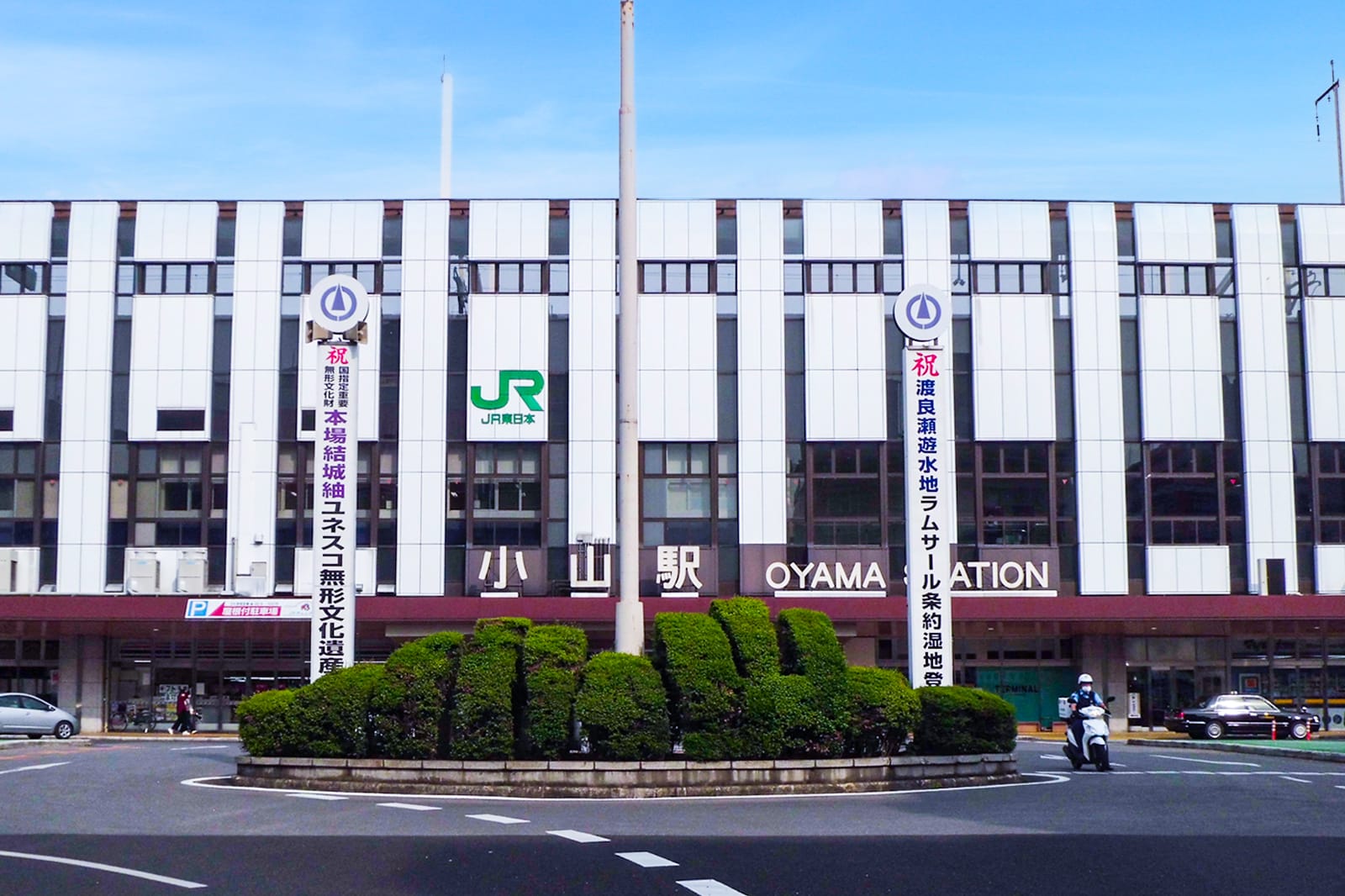 晴れた日の小山駅正面玄関、看板、横断幕、刈り込まれた生垣。.