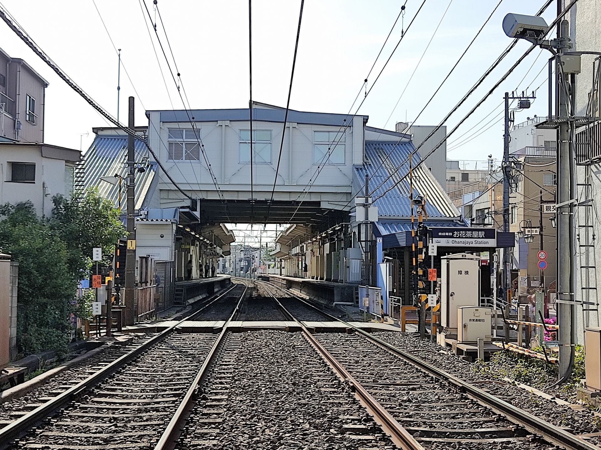 快晴の日、屋根付き歩道橋に向かう線路と、ホームや近隣の建物に囲まれた鉄道駅の眺め。.