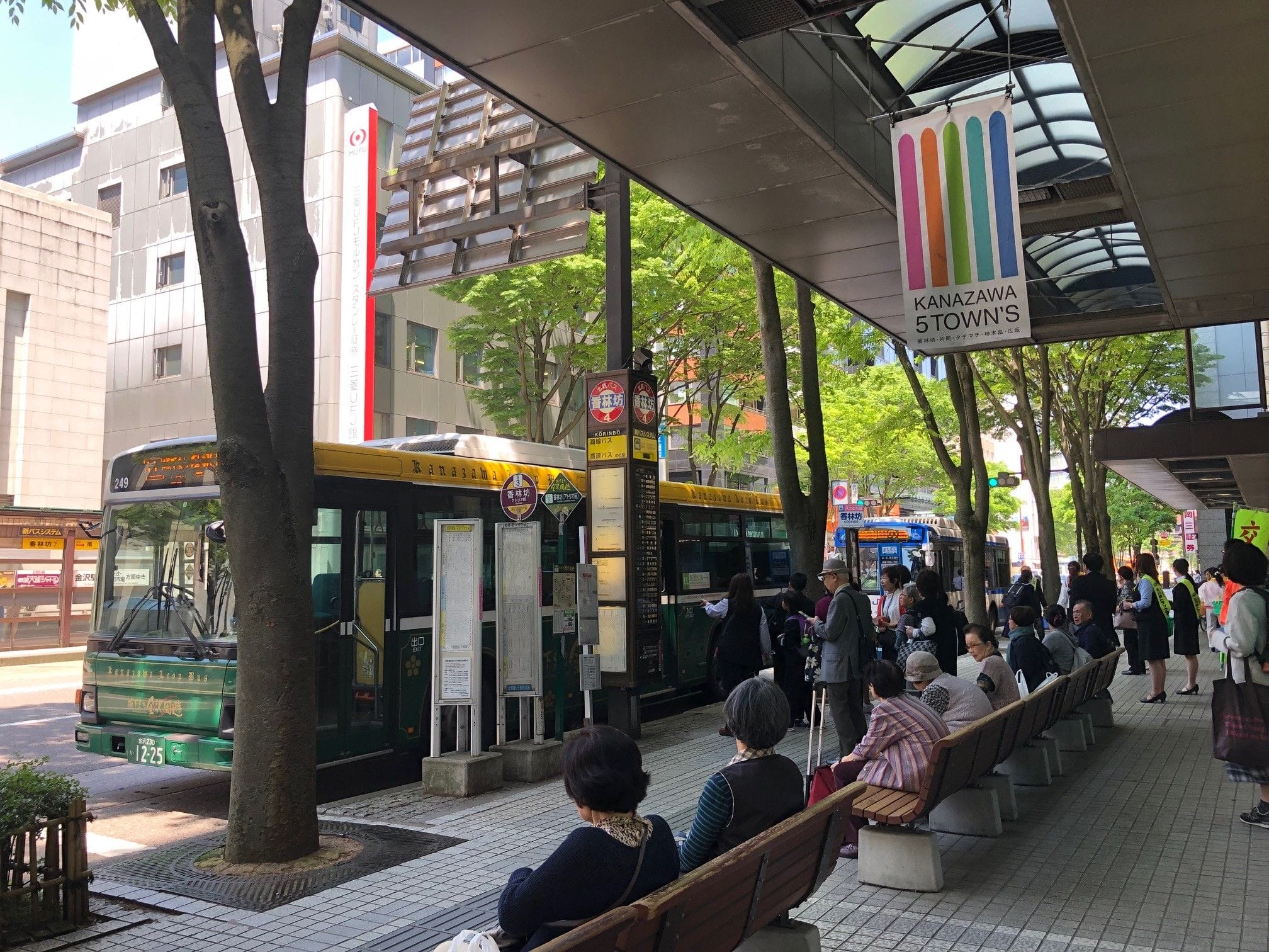 緑色のバスが停車し、もう1台のバスが近づいてくる市街地のバス停で待つ人々。看板には "KANAZAWA 5TOWN'S "と書かれている。街路樹が並ぶ。.