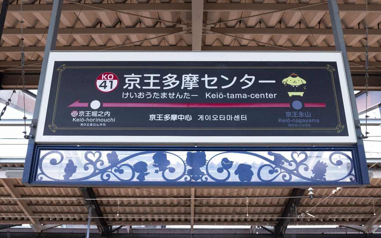 京王線の駅にある京王多摩センターへの案内看板。日本語、英語、韓国語、そして駅のマスコットのイラストが描かれている。.
