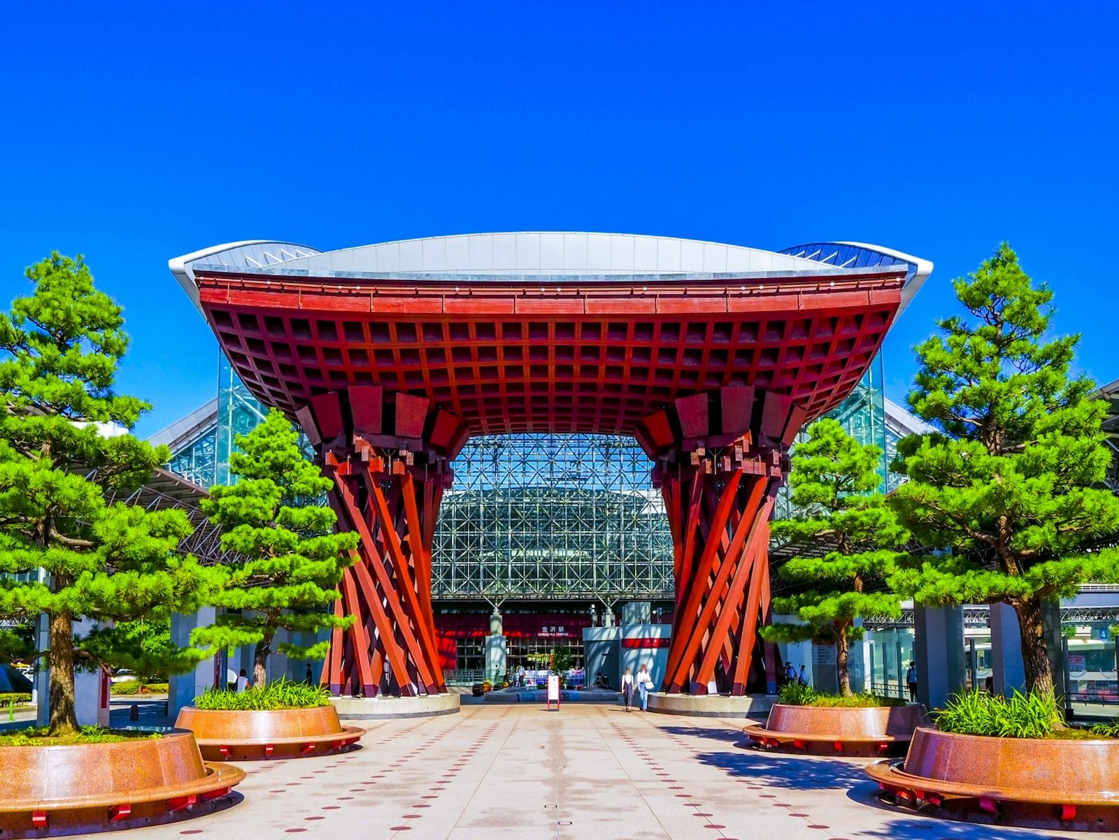澄み切った青空の下、緑の木々に挟まれたモダンなガラス張りの建物の前に、赤い柱の大きな木造の門構えが建っている。.