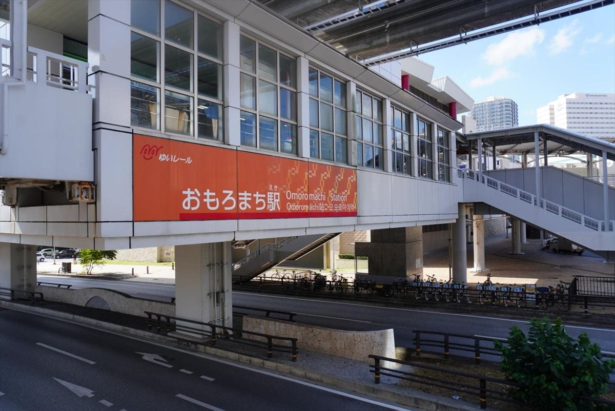 モノレールの高架駅。道路の上に日本語と英語で「おもろまち駅」と書かれた看板があり、その下に自転車が止まっている。.