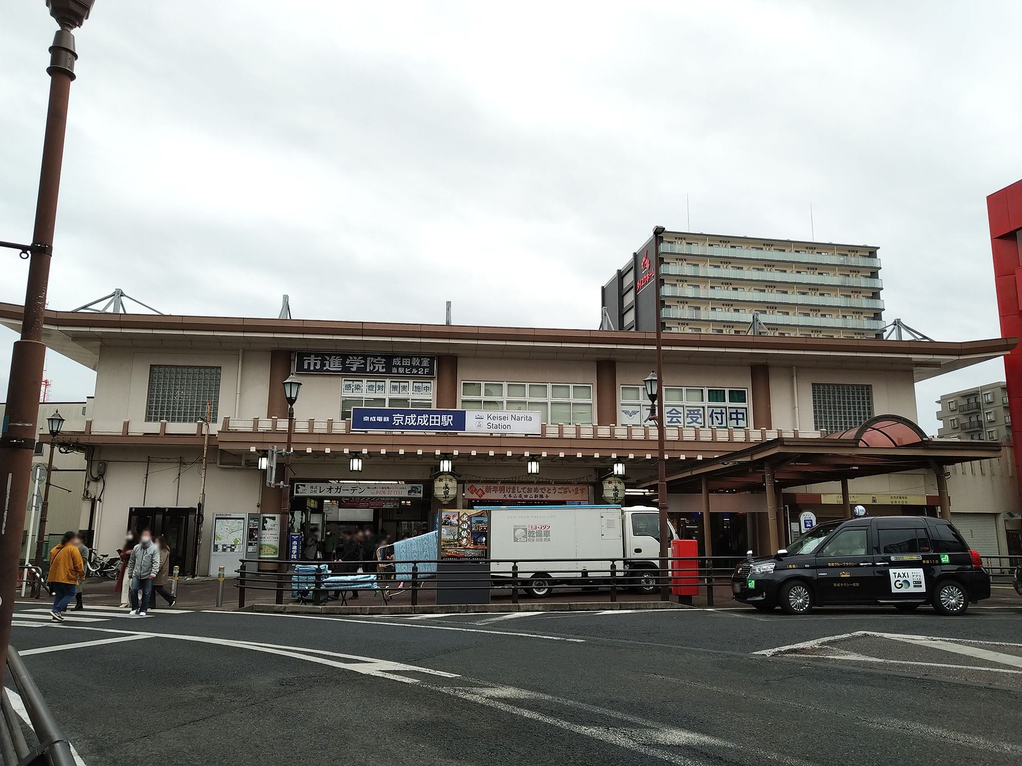 日本語の看板が掲げられた2階建ての駅舎、正面に停まるトラックとタクシー、そして曇りの日に近くを歩く人々。.