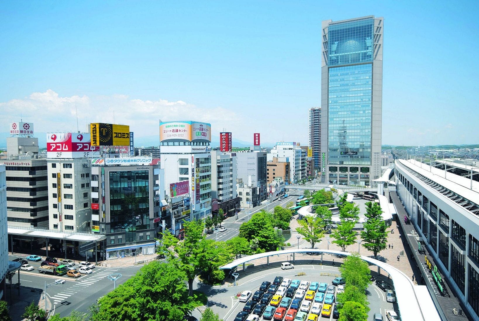 近代的な高層ビル、日本語の商業看板、車が行き交うカーブの多い道路、そして晴れた日の緑の木々。.