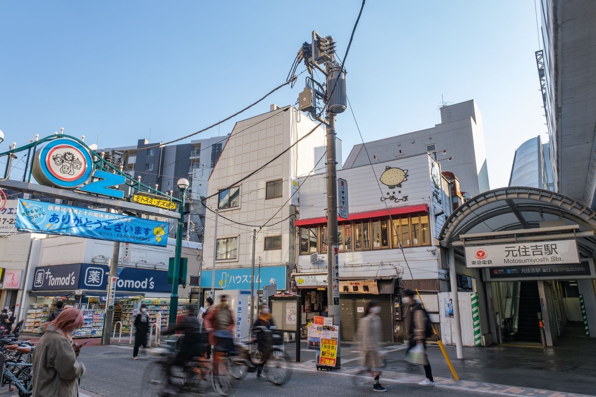 元越谷駅前の街並み。背景には歩行者や自転車、商店、看板、様々な建物が見える。.