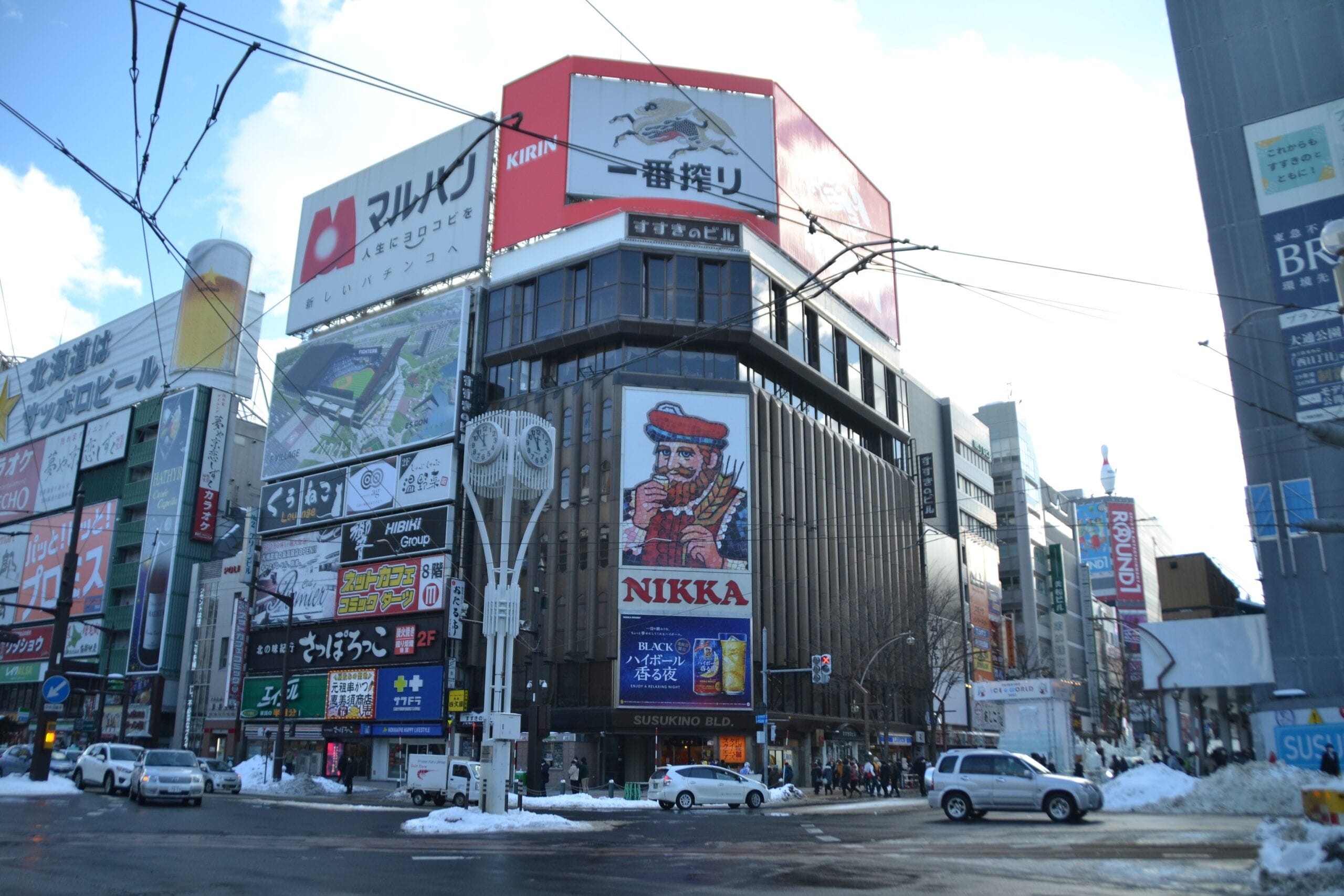 大きなビル、カラフルな看板、そして赤い帽子をかぶった男性が目立つニッカウヰスキーの広告。地面には雪が積もっている。.