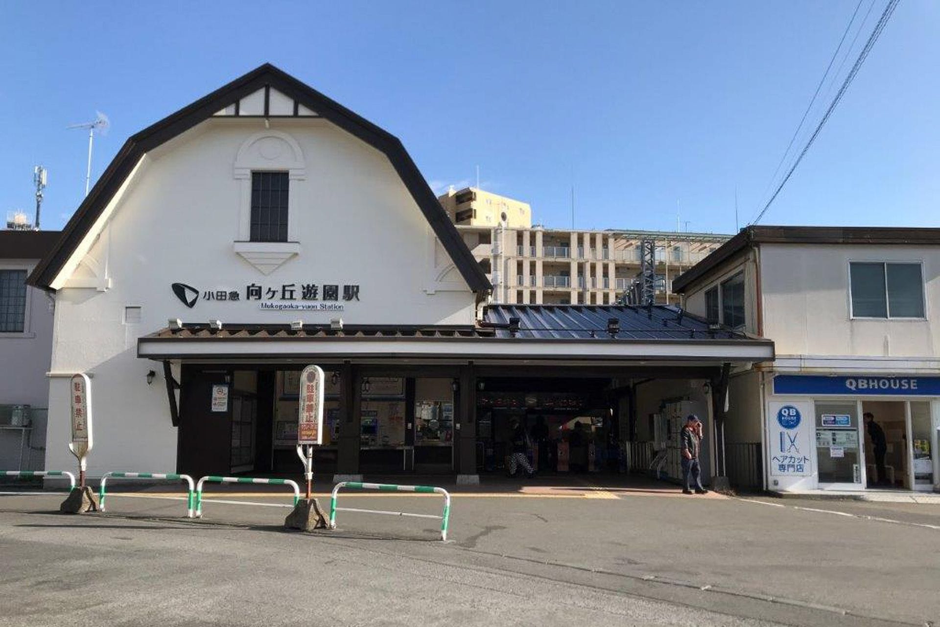 晴れた日の向ヶ丘遊園駅正面。入口付近に数人の人影と周囲の建物が見える。.