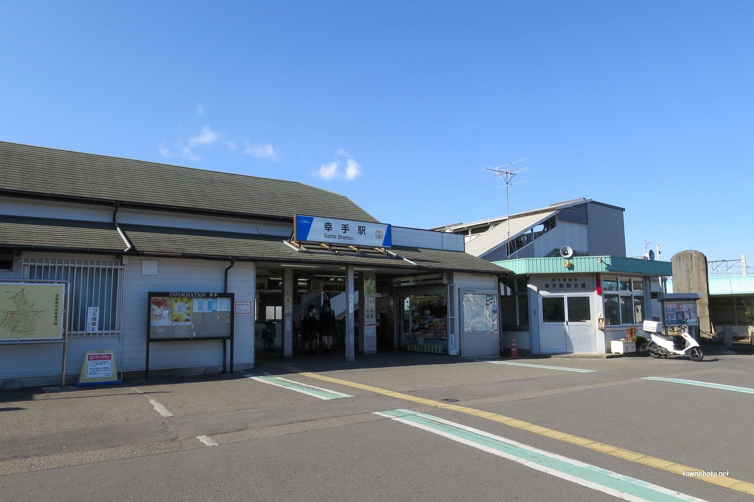 晴天の下、青い看板、券売機、高架歩道が隣接する小さな駅舎の外観。.