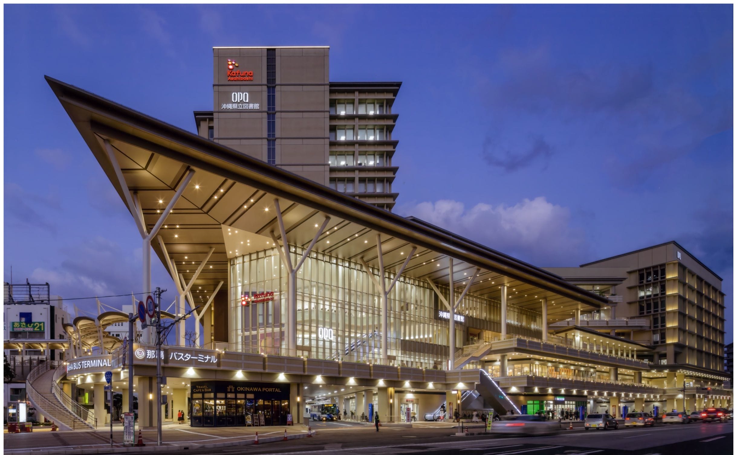 大きなガラス張りのファサードと角度のついた屋根を持つ近代的な駅舎が、夕暮れ時にライトアップされる。.