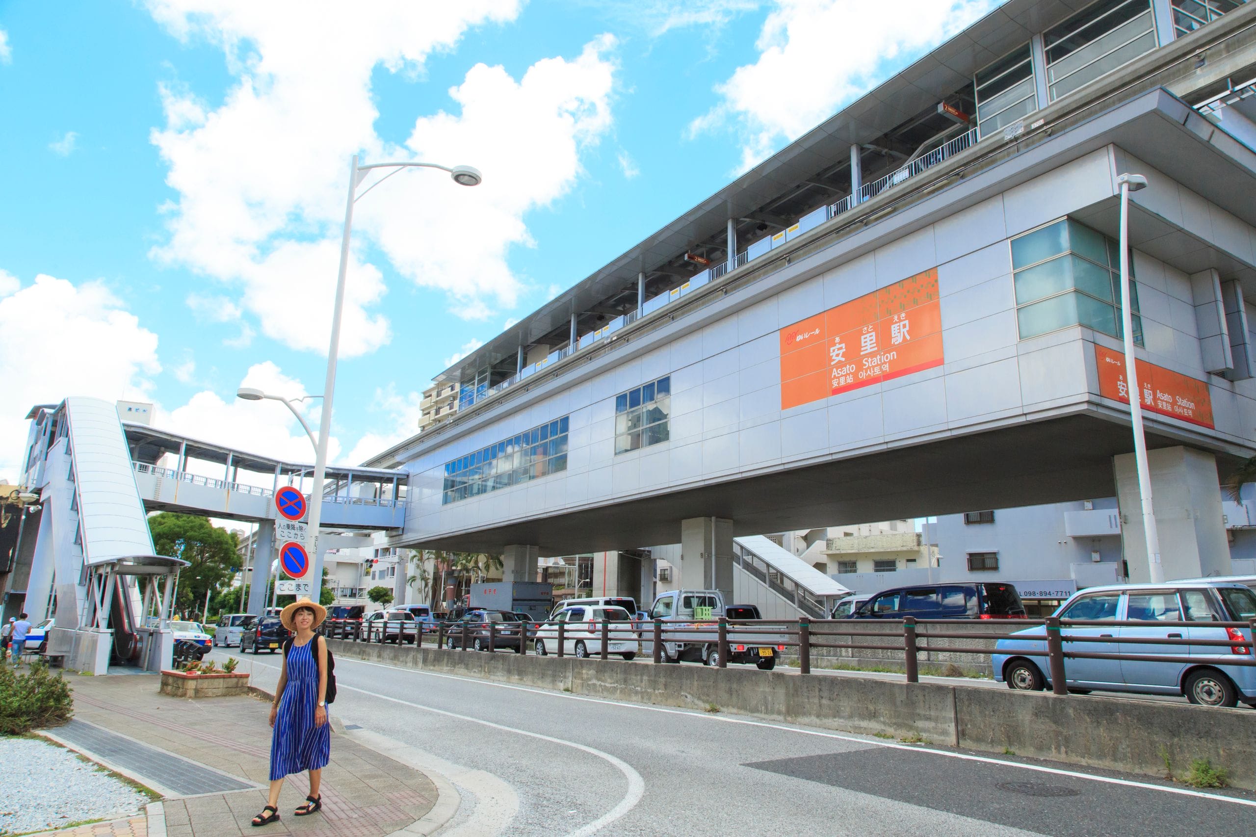 青いワンピースに帽子をかぶった女性が、下に車が駐車している近代的な高架鉄道駅近くの歩道に立っている。.