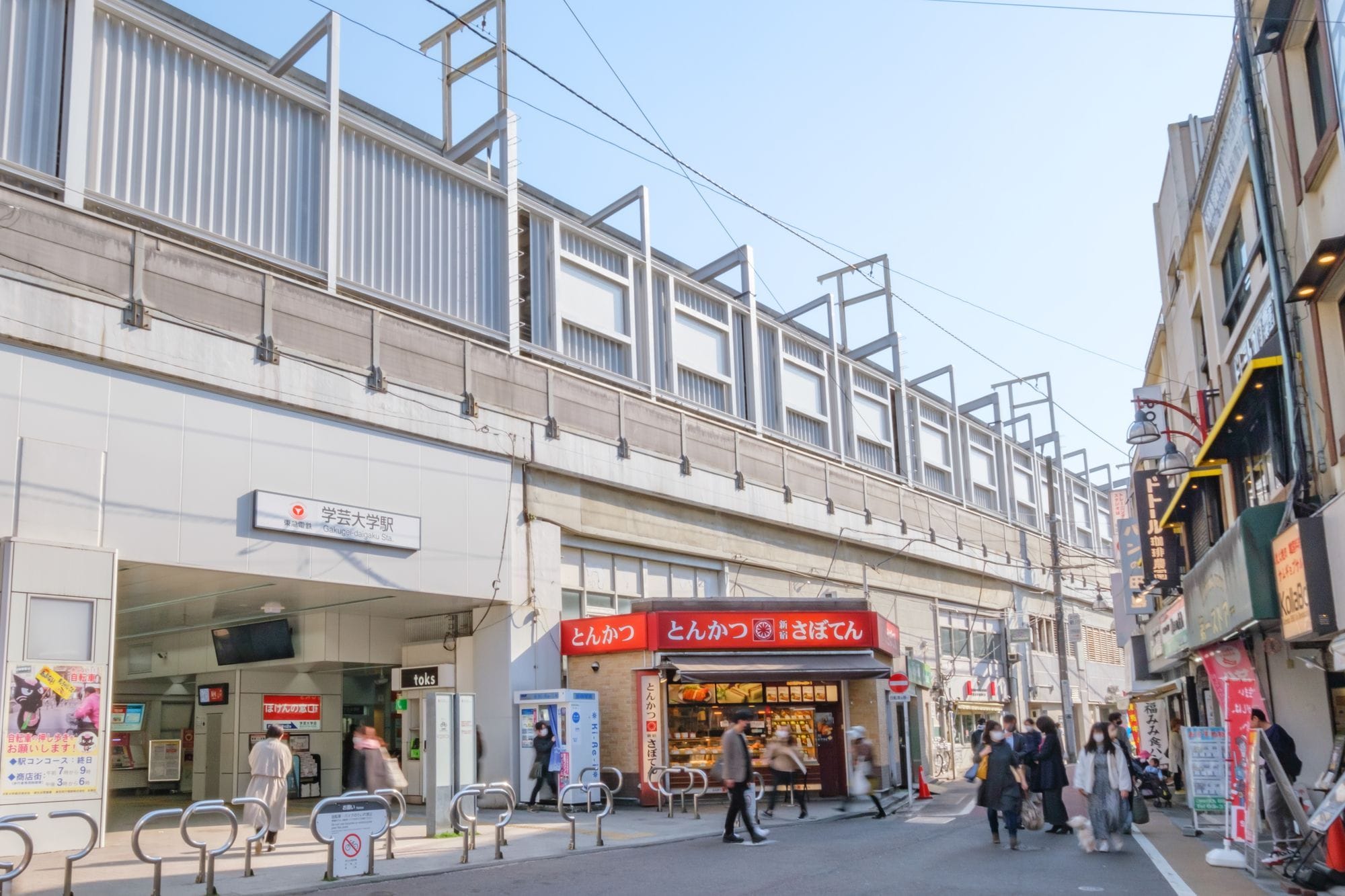 快晴の空の下、歩行者、自動販売機、店舗が並ぶ日本の駅入り口のストリートビュー。.