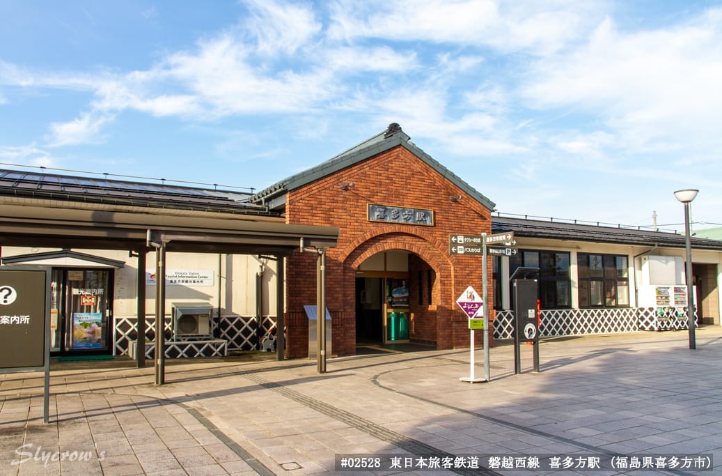 青空の下、レンガ造りの小さな駅の外観。分かりやすい看板、券売機、前面の舗装されたエリアがある。.