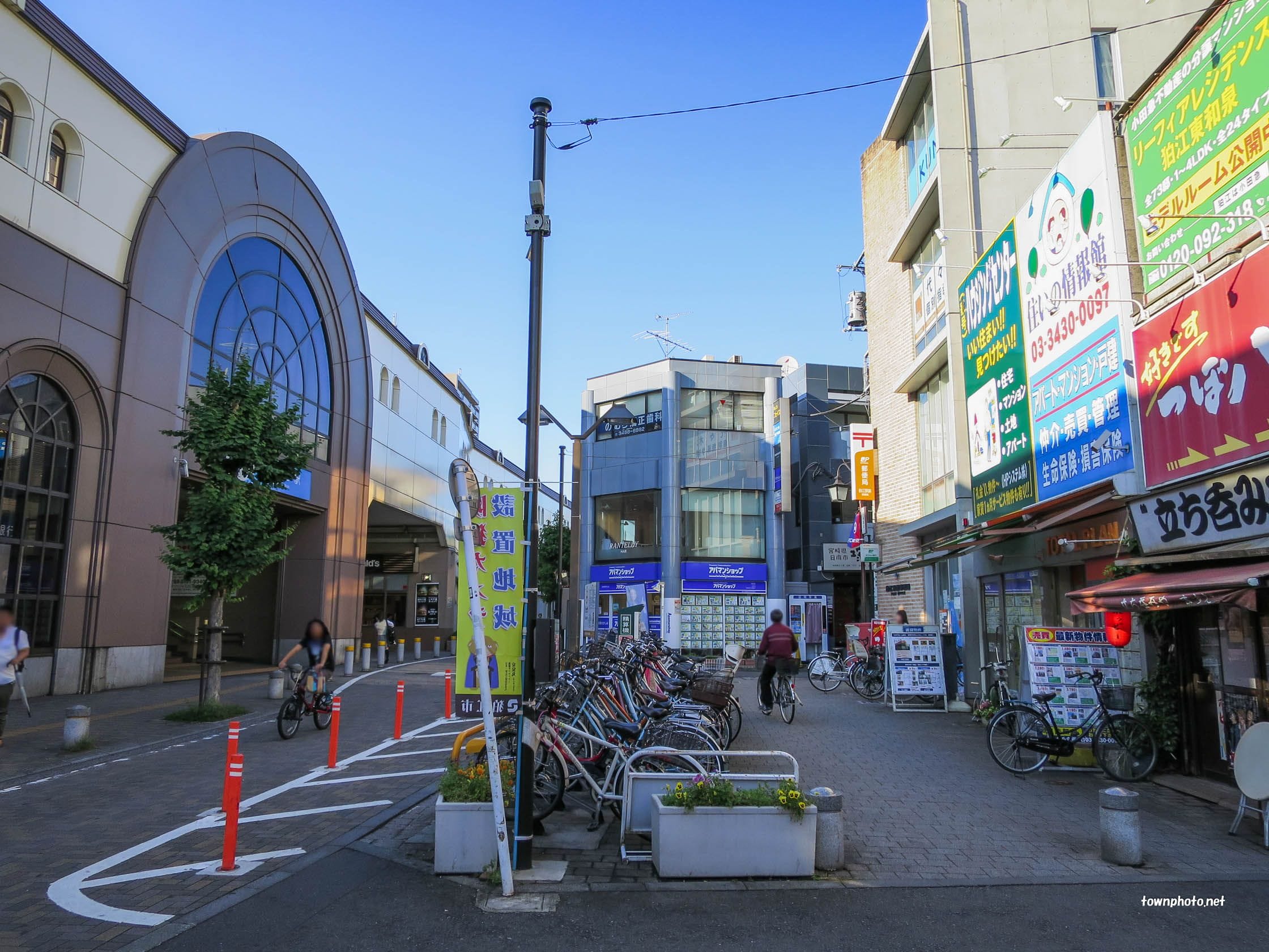 駐輪した自転車、散歩やサイクリングをする人々、商業ビル、日本語の看板、左側のモダンなアーチ型の入り口。.