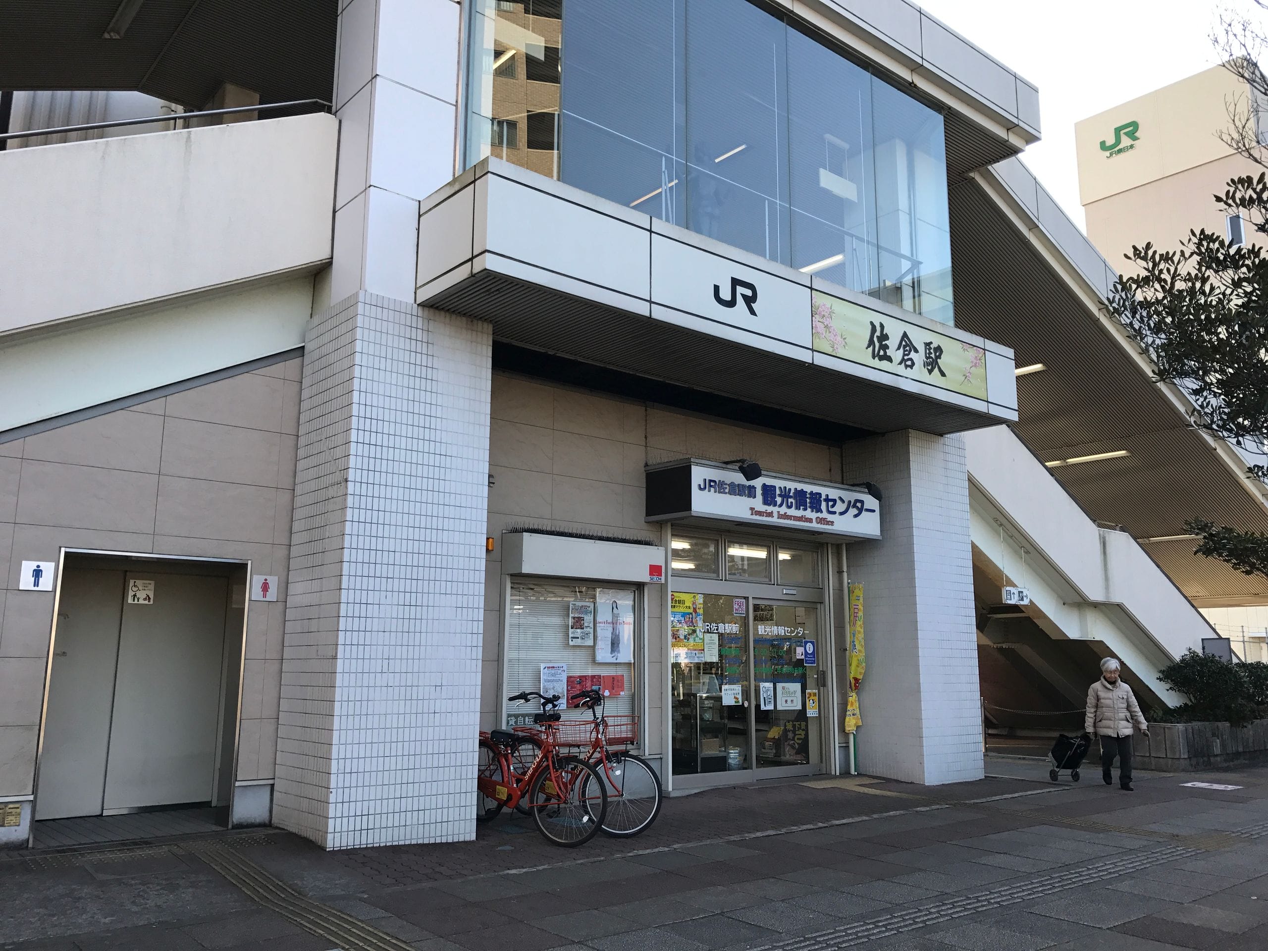 外に自転車が駐輪され、荷物を持って歩く女性、左手に公衆トイレの看板が見えるJRの駅入口外観。.