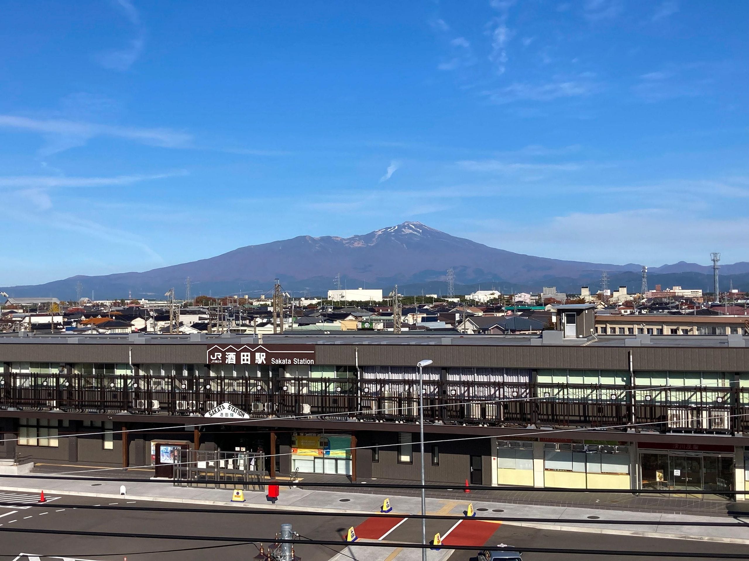 日本の榊駅の駅舎。手前には街並みが広がり、背景には澄み切った青空の下、雪を被った大きな山がそびえ立っている。.