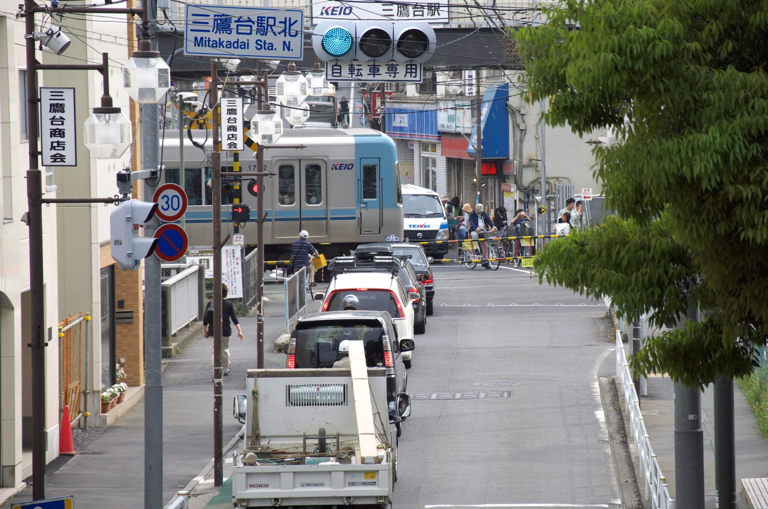 日本の通りで、青と白の電車が通り過ぎる中、踏切に車が列をなしている。通り沿いには標識や電柱、そして人々の姿が見える。.