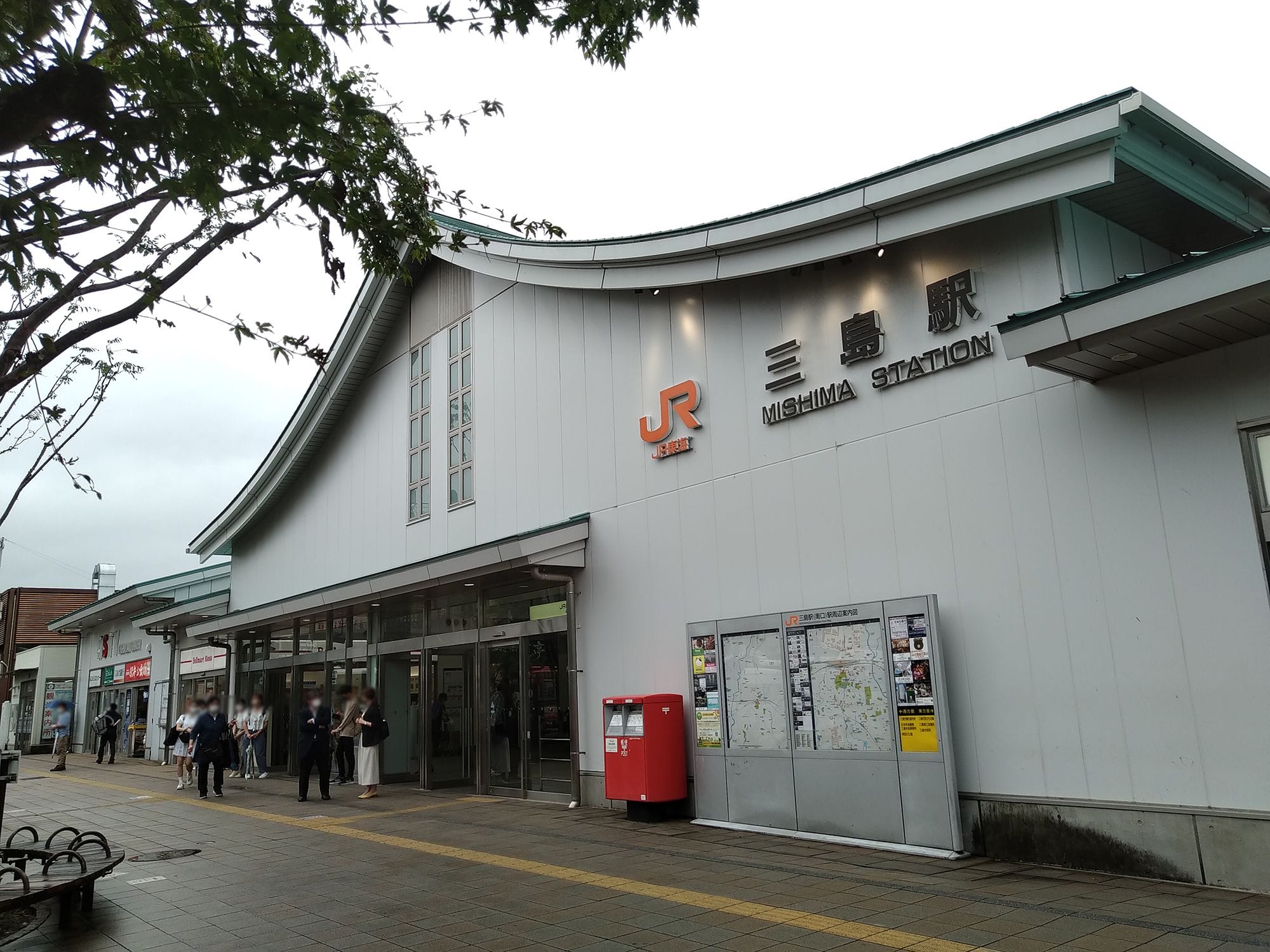 日本の三島駅の外観。駅の入口、日本語と英語の案内表示、地図、赤い郵便ポストが写っており、その周辺には数人の人がいます。.