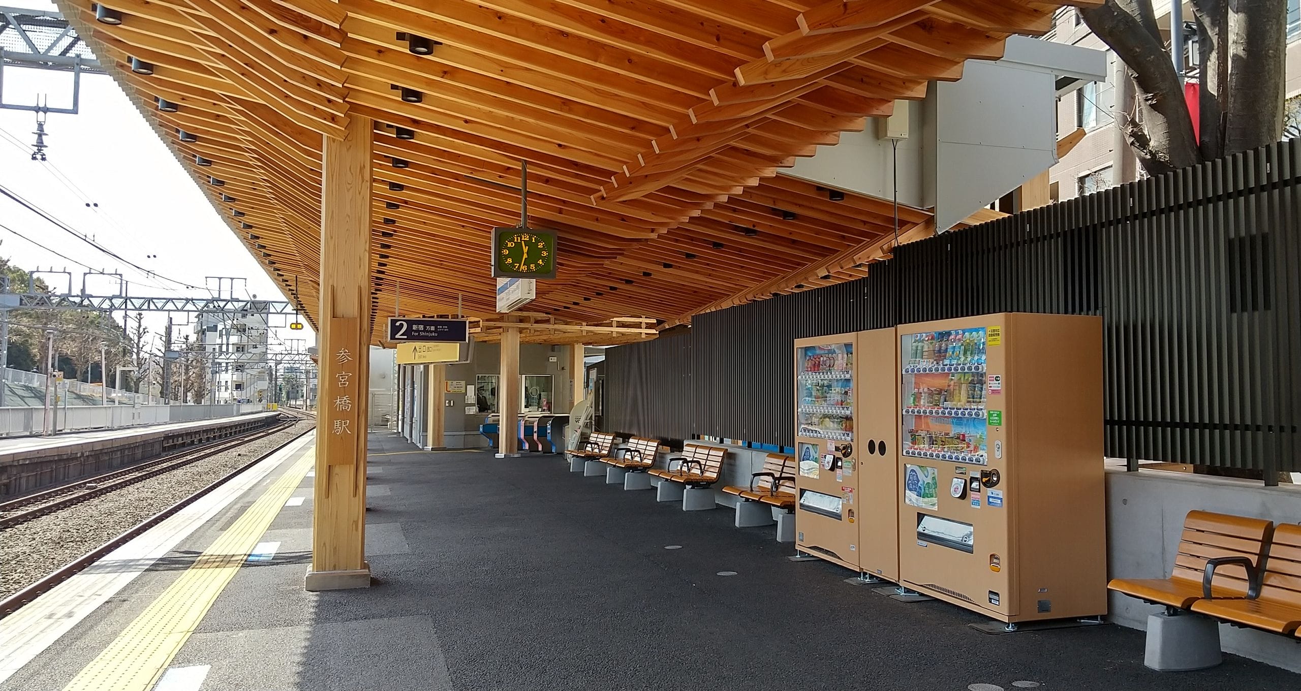 木製の屋根、自動販売機、ベンチ、時計、案内板がある駅のホーム。人は誰もいない。左側には線路が走っている。.