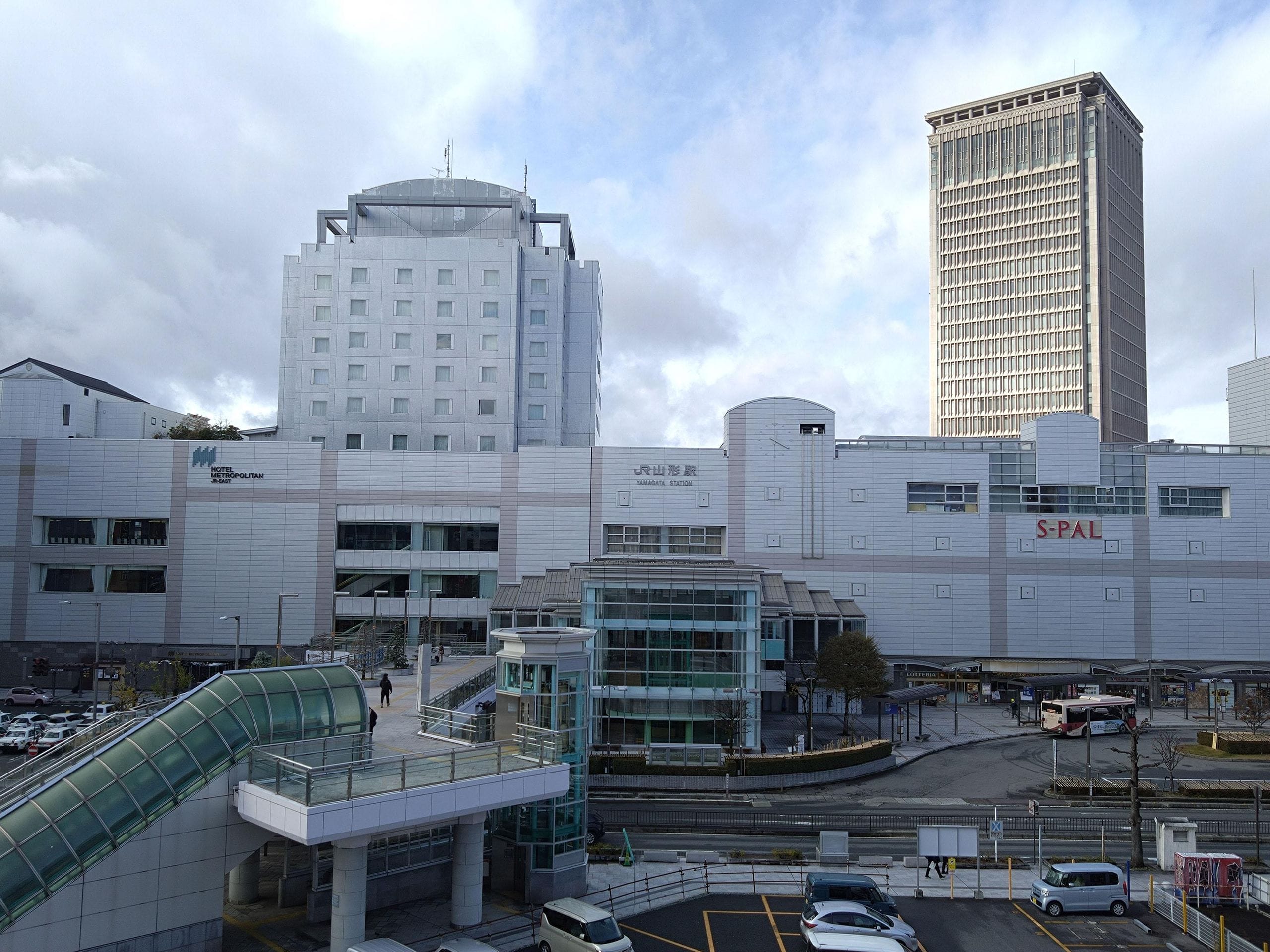 「S-PAL」の看板が掲げられた大きな白い建物と、その隣にそびえる高層オフィスビルが写った街並み。手前には車やバス、歩行者が映っている。.