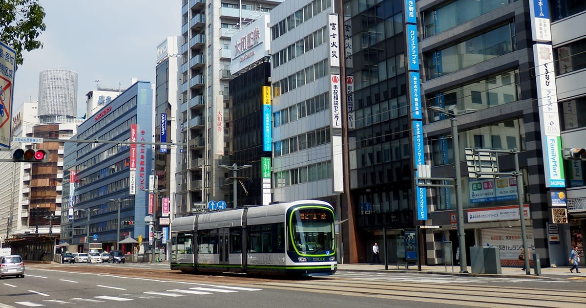 近代的な路面電車が、日本語の看板が掲げられた中層のオフィスビルが立ち並ぶ街路の線路を走っている。.