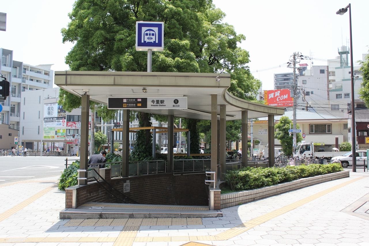緑に囲まれ、ビルを背景にした街路に隣接する、頭上に看板のある都市部の地下鉄駅の入口。.
