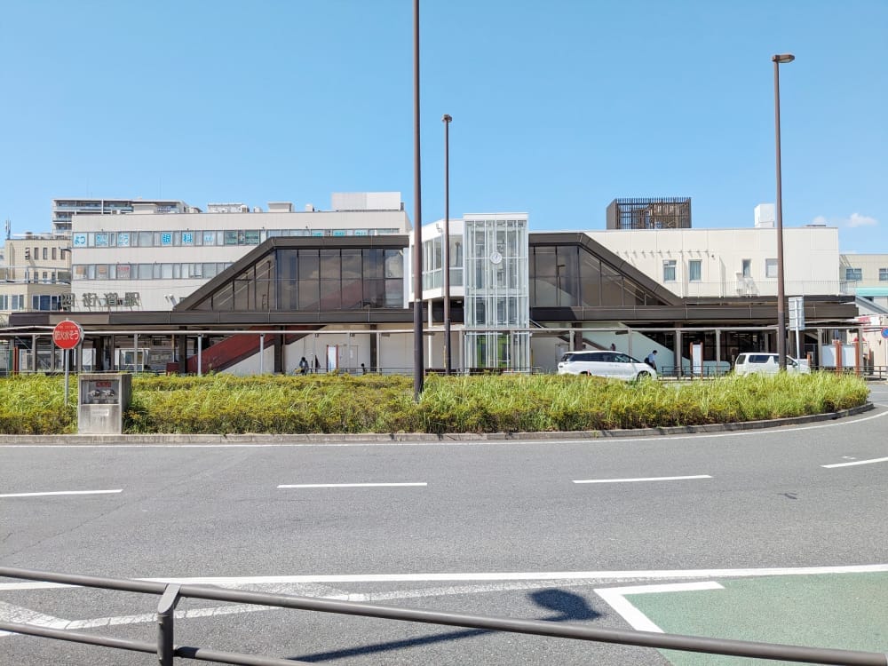 ガラス張りの歩道橋やエレベーターを備えたモダンな建物。外には白いバンが数台停まっている。手前には青空が広がり、停止標識が見える。.